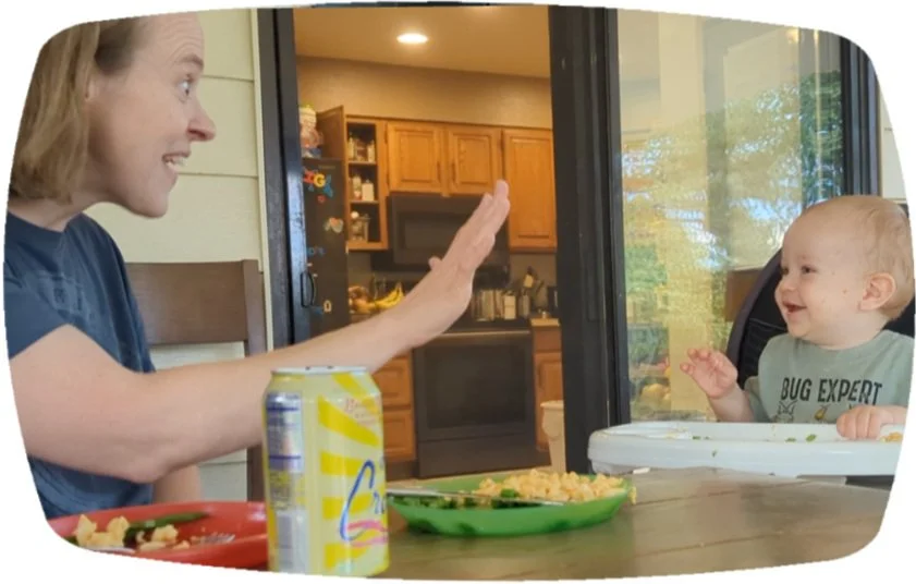 woman and baby at a table with food. Woman is signing "5" in ASL; baby is laughing from a high chair