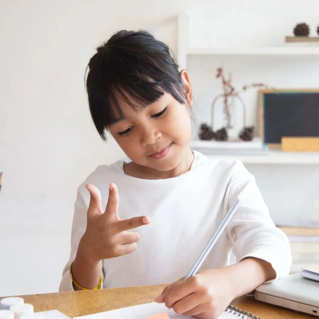 child with medium-dark skin in a white shirt, holding up thumb and two fingers (number 3 in ASL)
