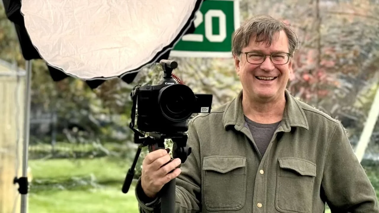 Scott Indermaur holding a camera on location with a softbox behind him during a video production setup.
