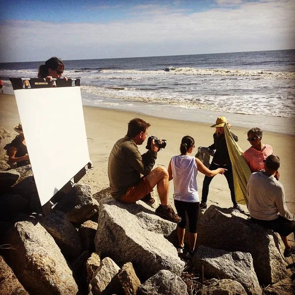 Behind-the-scenes beach video production with crew, camera operator, and lighting tools during an on-location shoot.