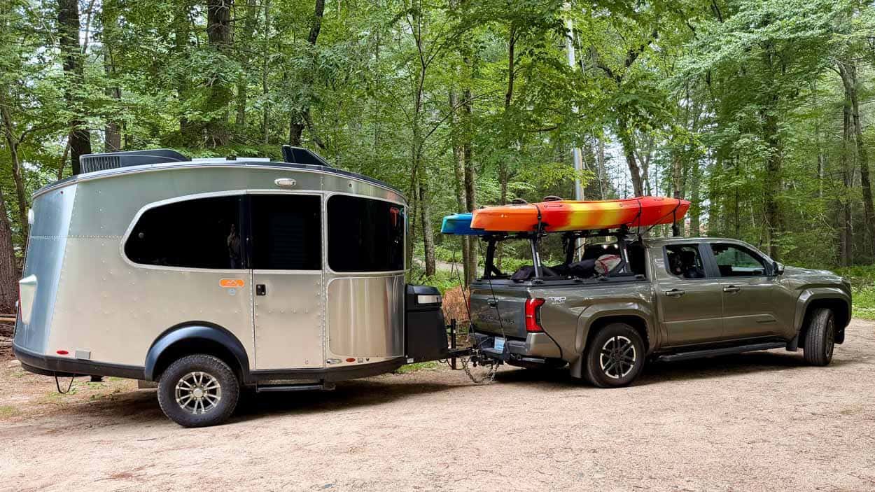 Toyota Tacoma towing an Airstream Basecamp with two kayaks mounted above the truck bed, parked on a wooded Rhode Island property.