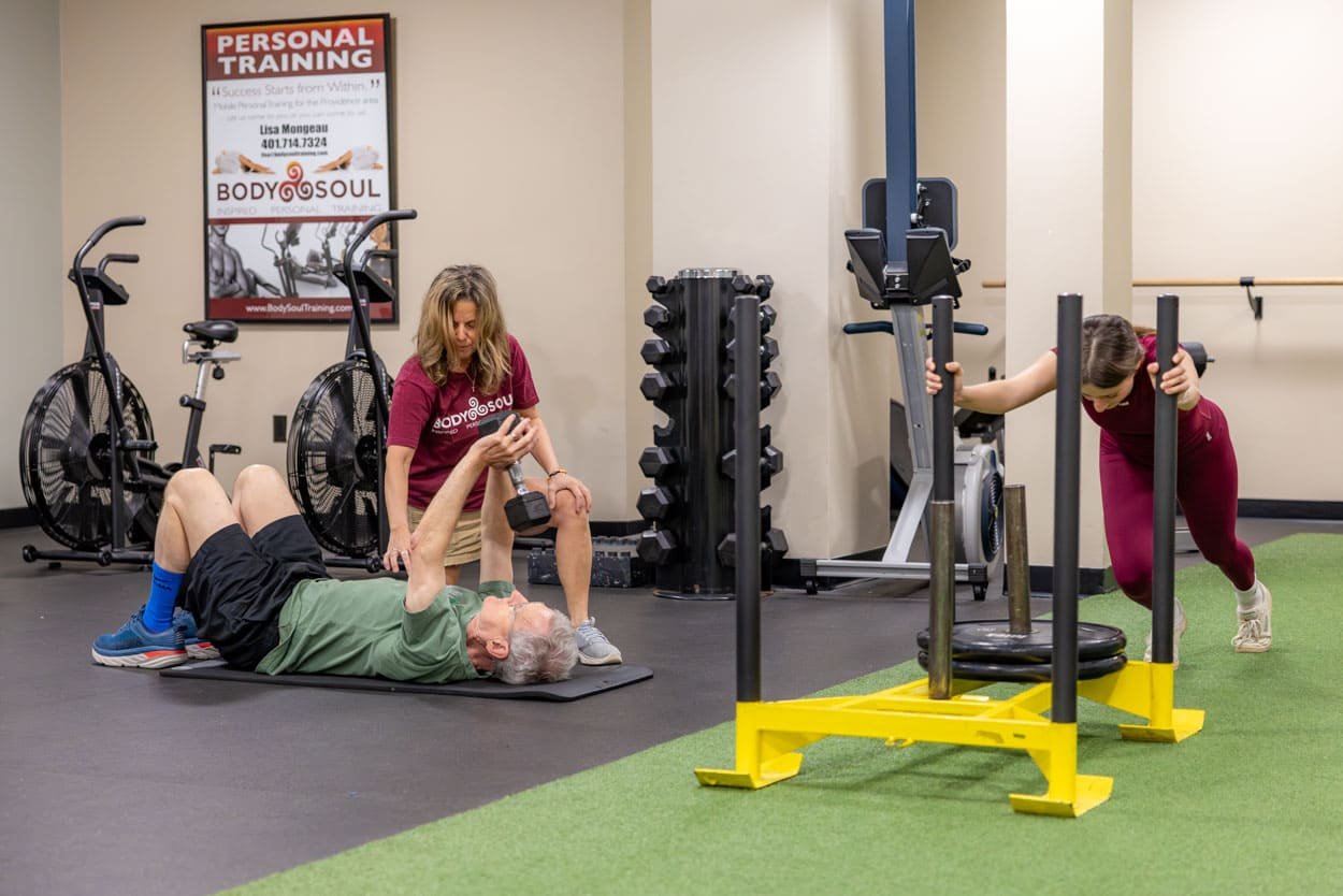 Body Soul Training trainer assisting a client with a floor exercise while another client performs a weighted sled exercise in the Providence studio.
