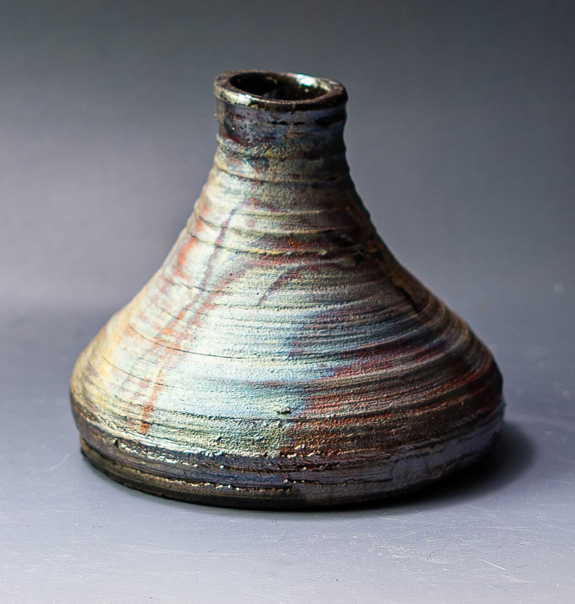 A small, textured ceramic pottery piece with an earthy, metallic glaze, conical shape, and narrow opening at the top, placed on a gray surface.