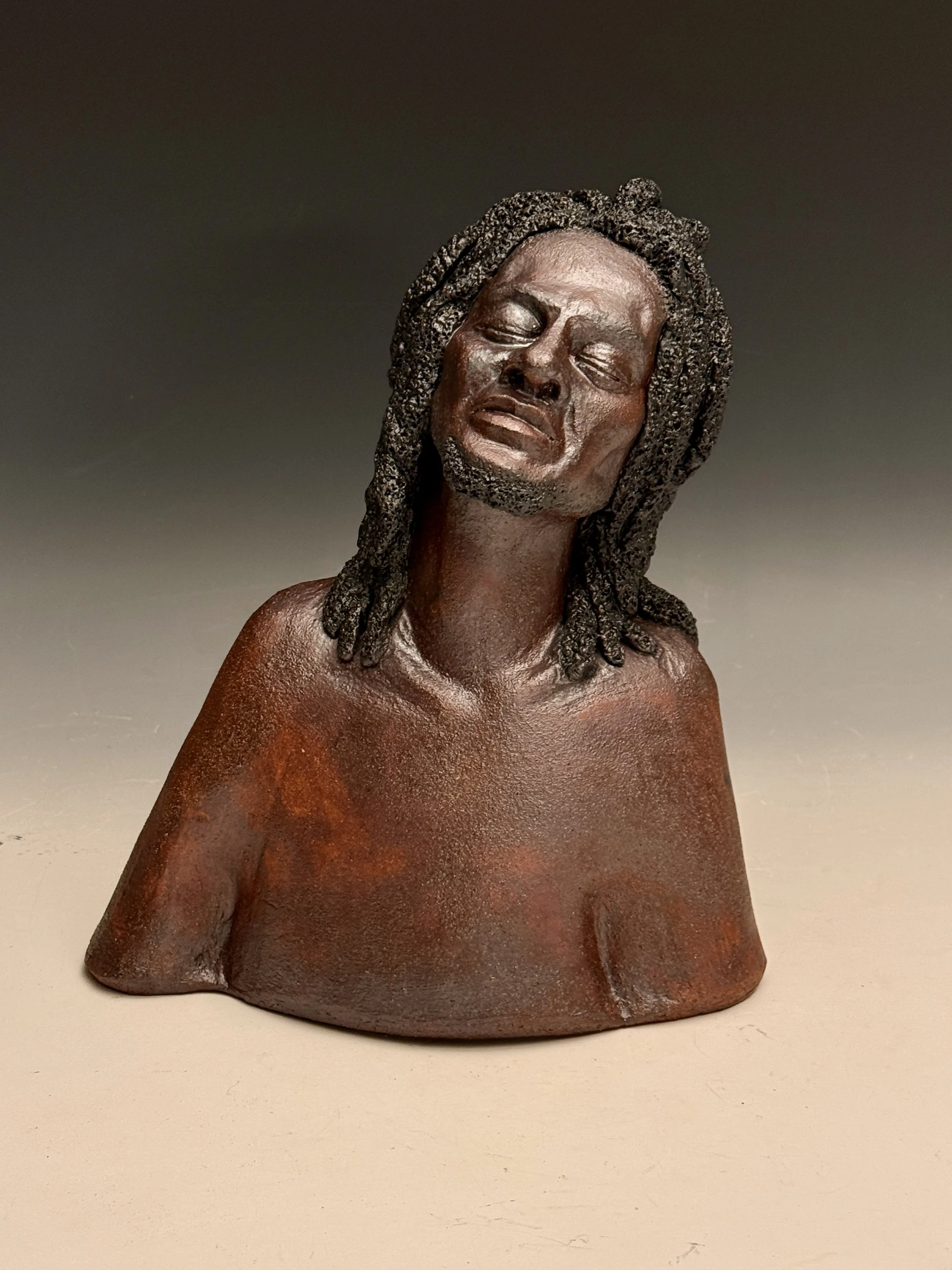 Bronze bust sculpture of a woman with her eyes closed and an expressive, emotional face, featuring detailed dreadlocks and a textured surface.