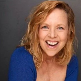 A woman with curly blonde hair wearing a blue top, smiling with arms crossed.