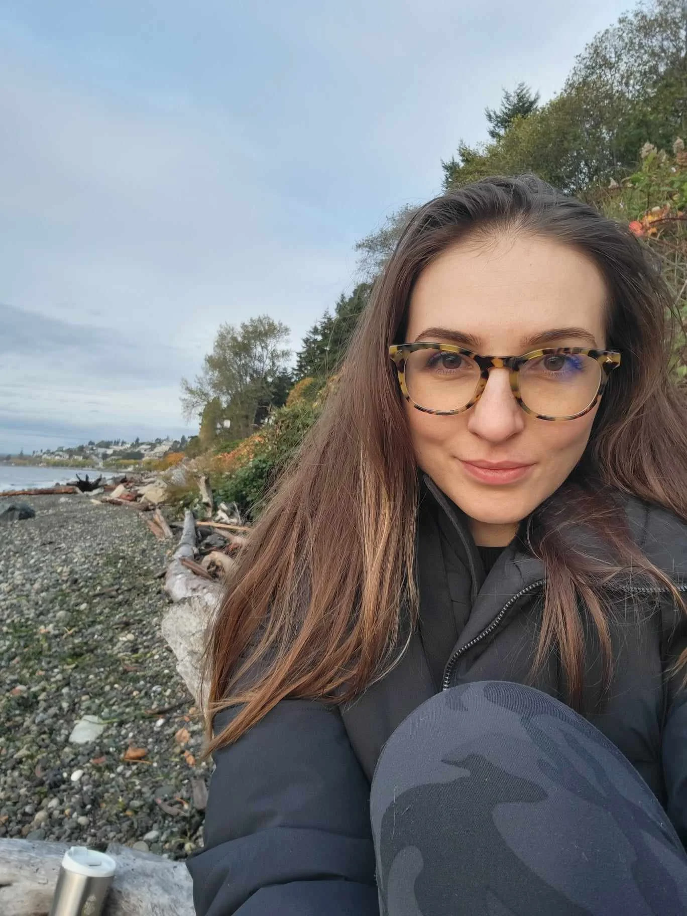 A woman with long brown hair and glasses sitting on a rocky beach, looking at the camera, with trees and houses in the background.