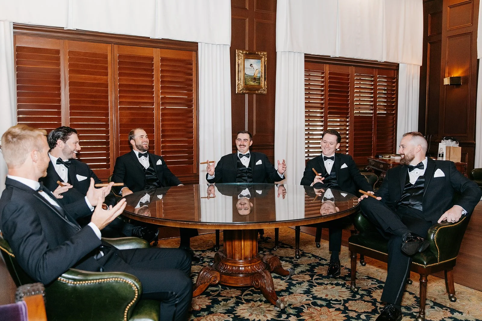 Six men in tuxedos sitting around a polished wooden oval table in a formal room with wooden shutters and white curtains, engaging in conversation and smiling.