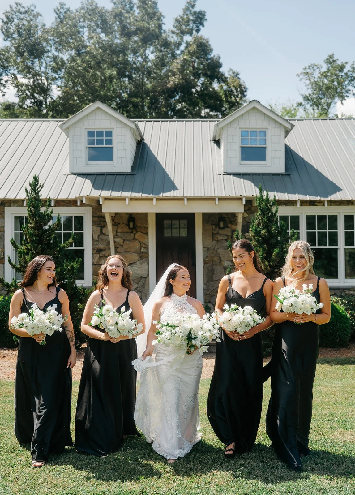 Bride and bridesmaids walking outside in front of a stone house, smiling and holding white bouquets on a sunny day.