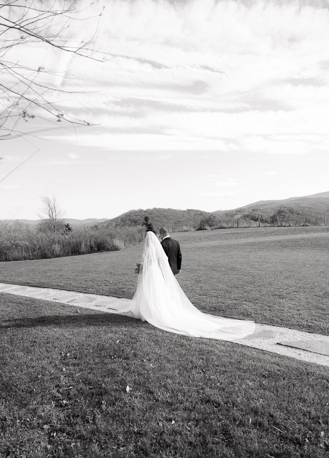 A bride and groom walking hand in hand on a sidewalk in an open grassy field with mountains in the background, some trees, and a partly cloudy sky.
