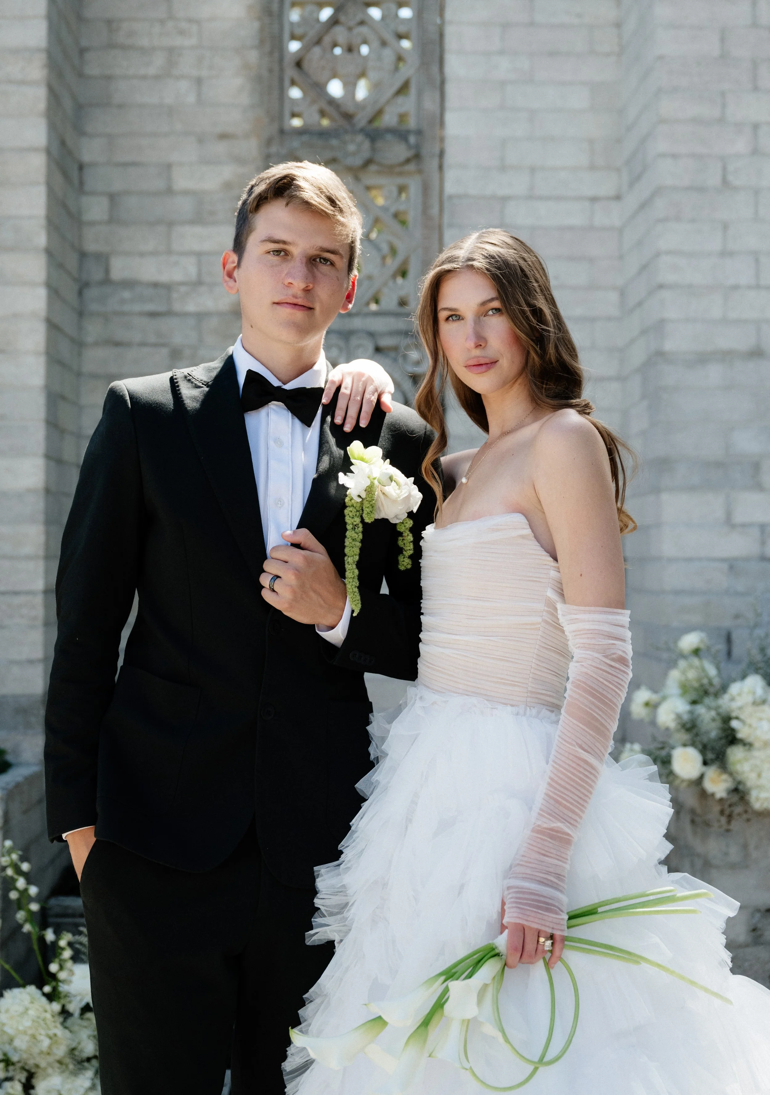 A bride and groom stand close together outdoors, wearing wedding attire, with flowers and decorative background behind them.
