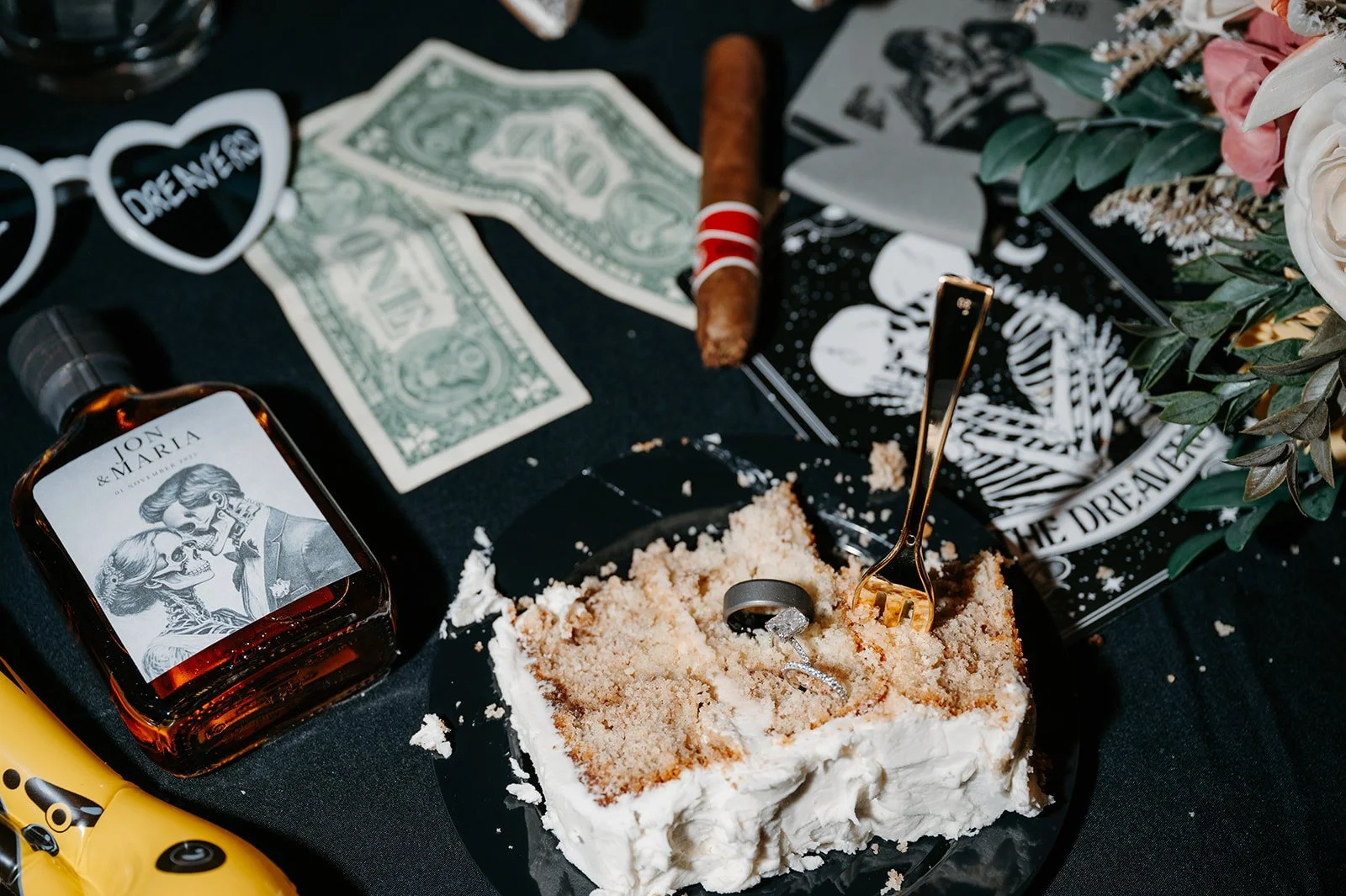 A partially eaten cake on a black plate with a gold fork, a bottle of liquor with a skeleton and couple label, dollar bills, a cigar, and decorative items including a heart-shaped glasses that says 'Dreamer' and a floral arrangement.
