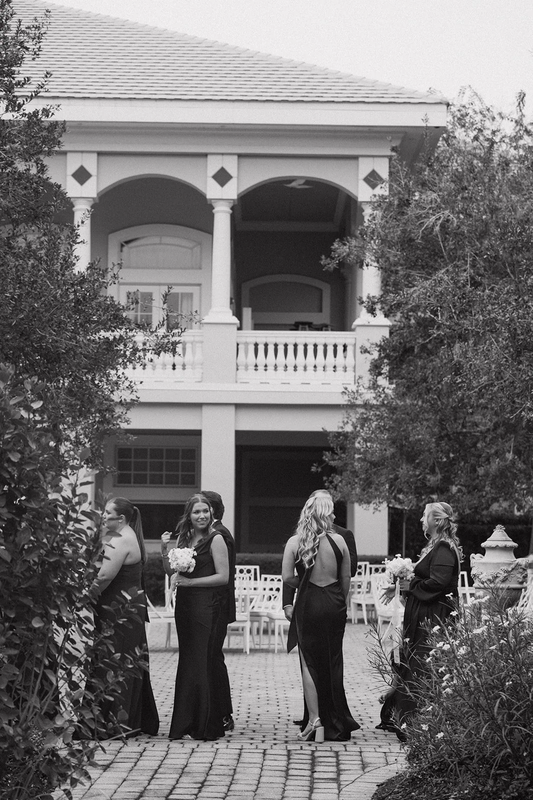 Women dressed in formal black gowns standing outdoors near a large house with a balcony, some holding bouquets of flowers.