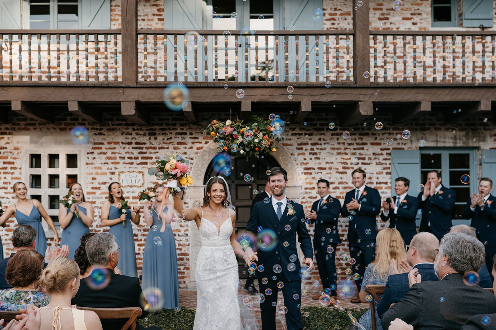 A newlywed couple is walking down the aisle, surrounded by friends and family, during a wedding celebration outside a rustic brick building, with soap bubbles floating in the air.