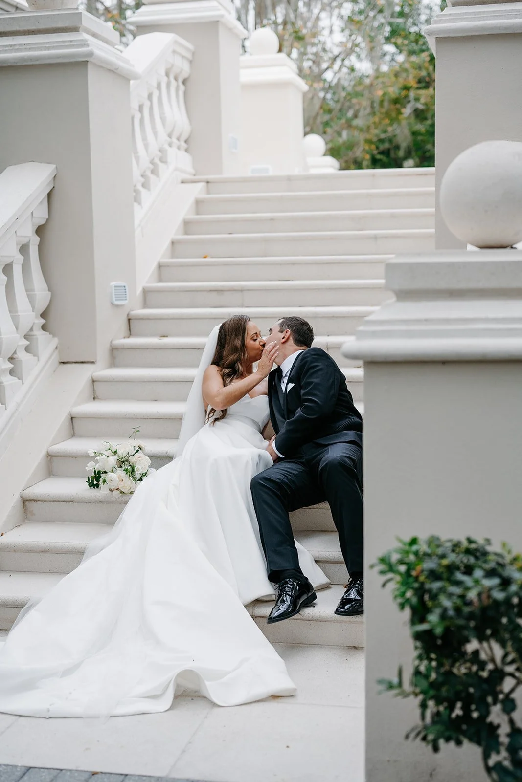 A bride and groom sit on white marble stairs; the bride is in a white wedding gown with a long train, holding a bouquet of white flowers, and the groom is in a black tuxedo; they kiss.