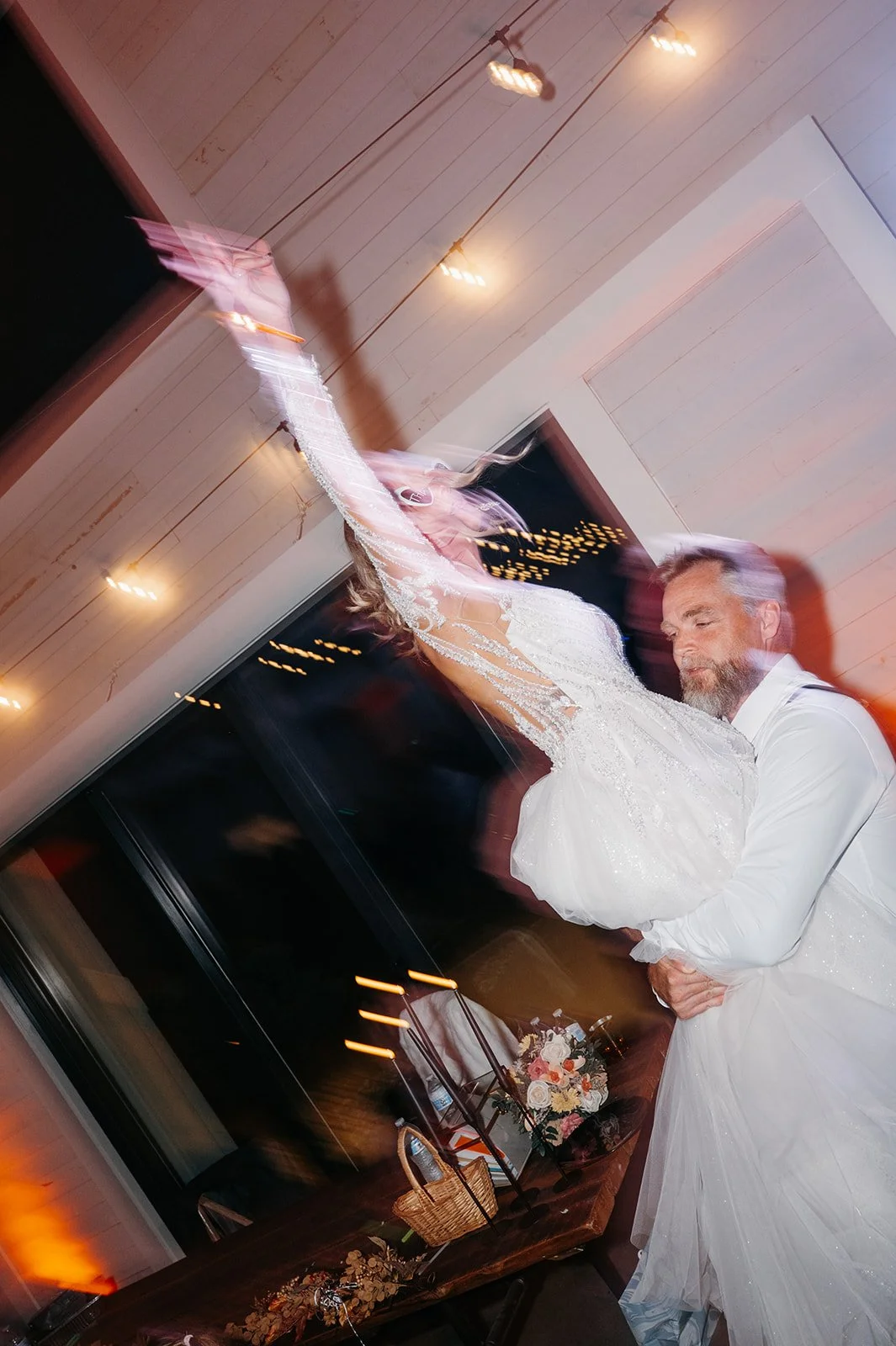 A bride and groom sharing their first dance at a wedding reception. The bride is wearing a white wedding gown and the groom is dressed in a white shirt. The photo has a motion blur effect.