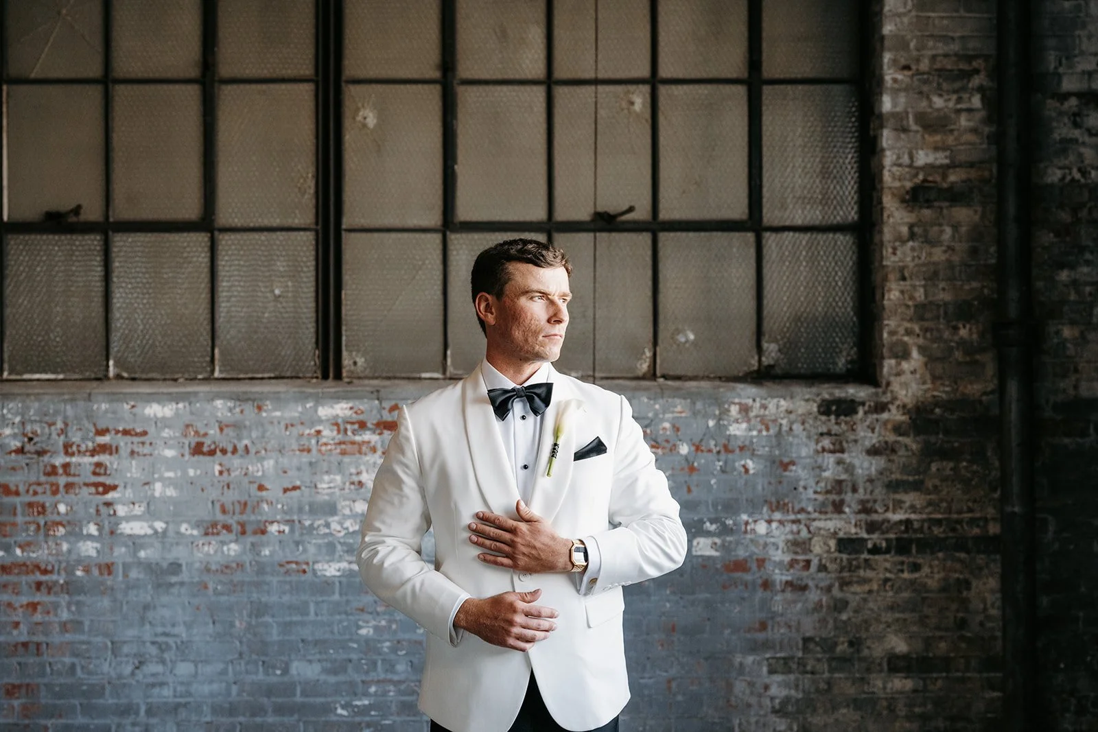 A man in a white tuxedo with a black bow tie and watch, standing in front of a rustic brick wall with large industrial-style windows, looking to the side.