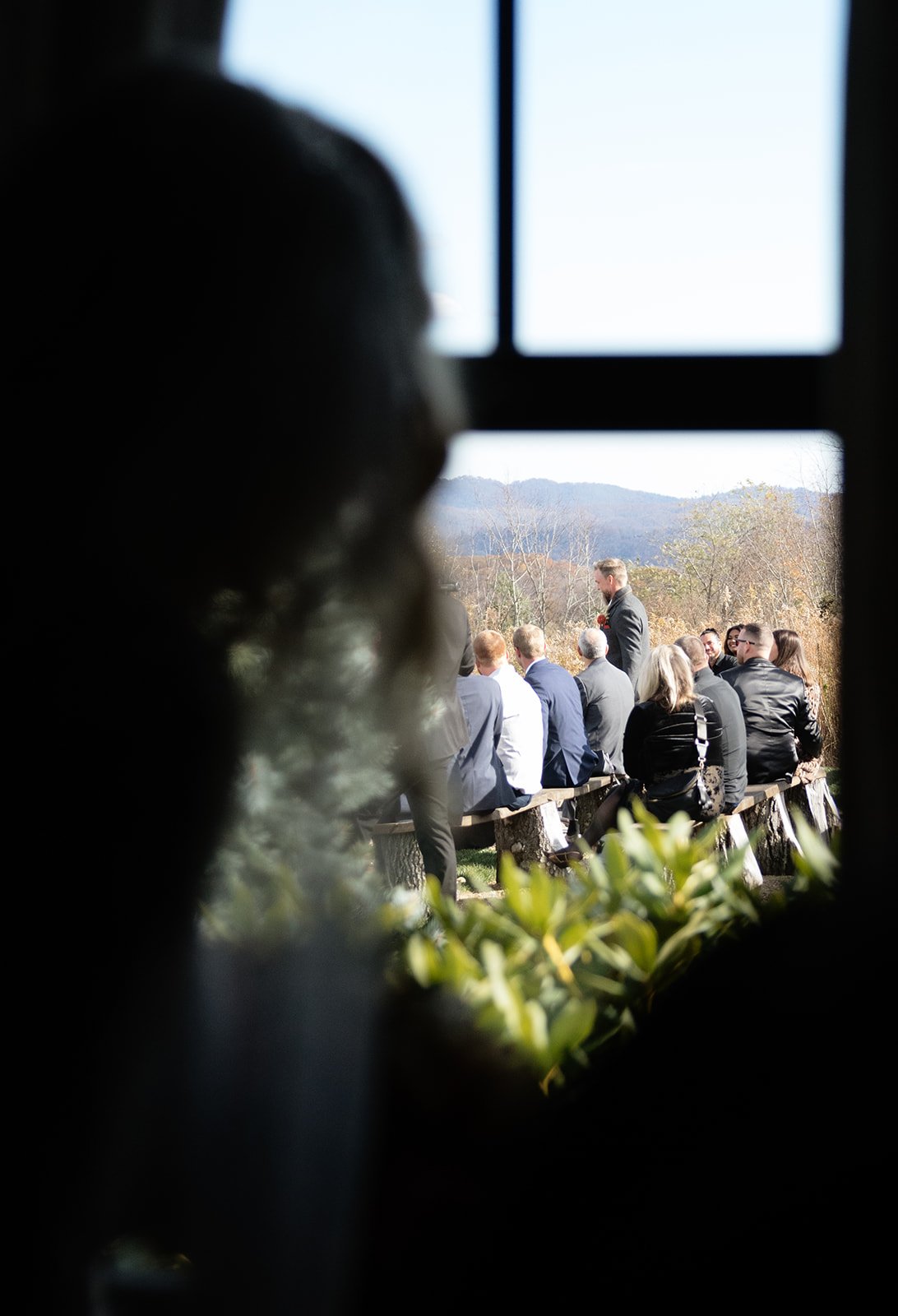 View through a window of a wedding ceremony outdoors with guests seated on benches and mountains in the background.