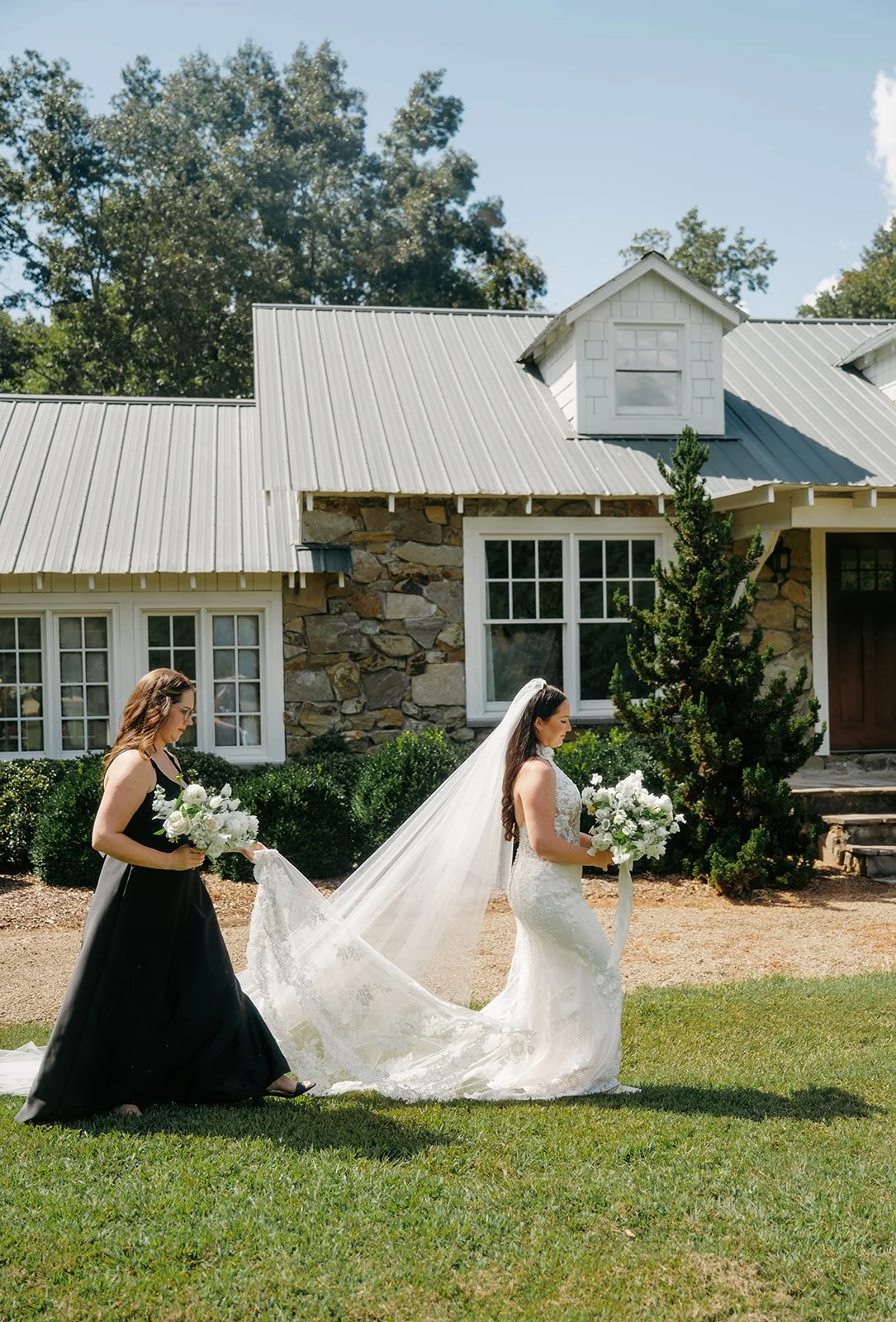 A bride in a white wedding dress and veil holding a bouquet walks on grass in front of a stone house, while a woman in a black dress adjusts her train.