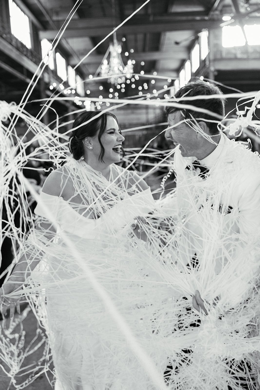 A bride and groom dancing together at their wedding reception, surrounded by white streamer decorations, smiling and laughing in a large, decorated indoor venue.
