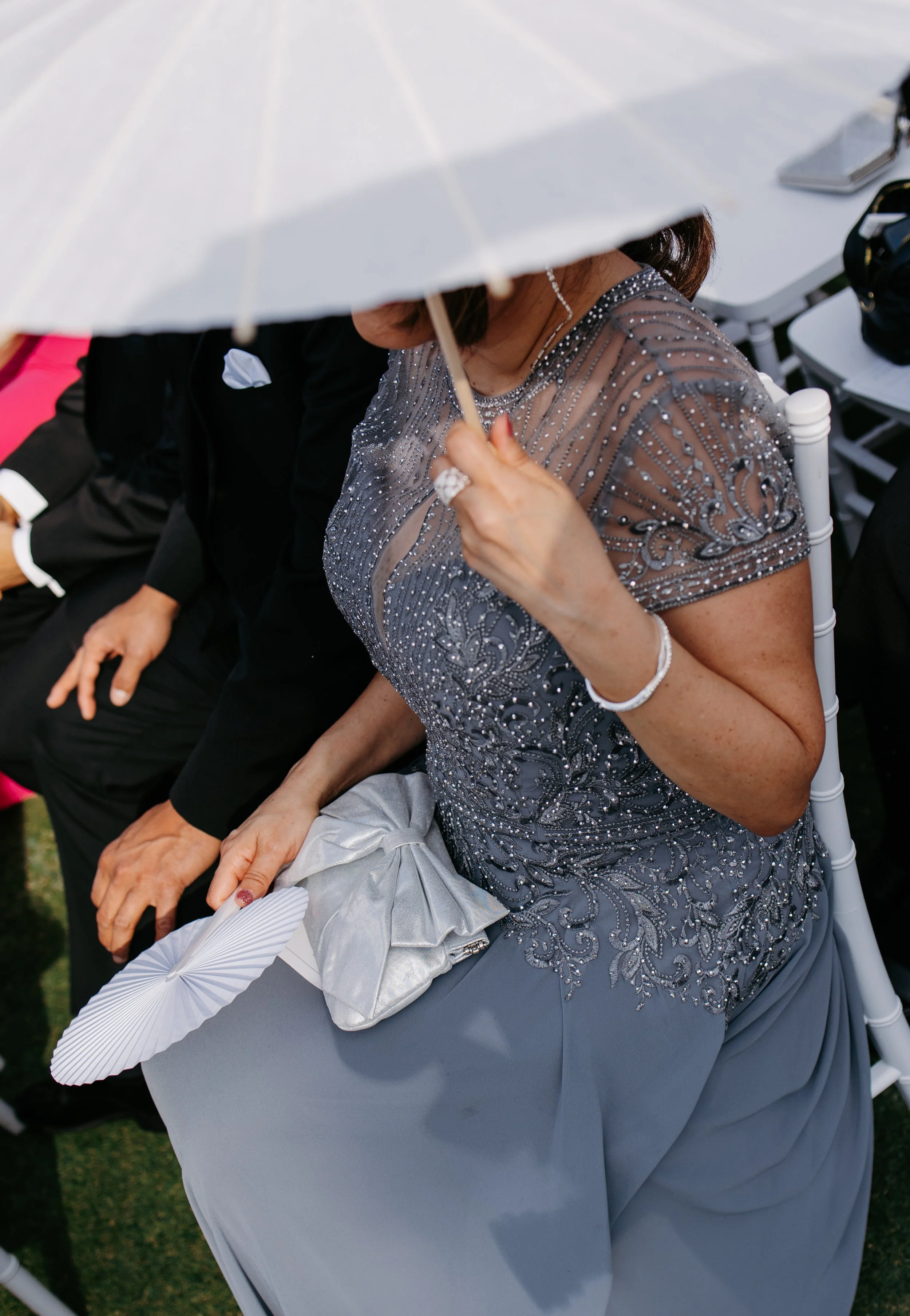 Woman in a grey, embellished dress sitting at a formal event, holding a silver clutch, with a white umbrella shading her.
