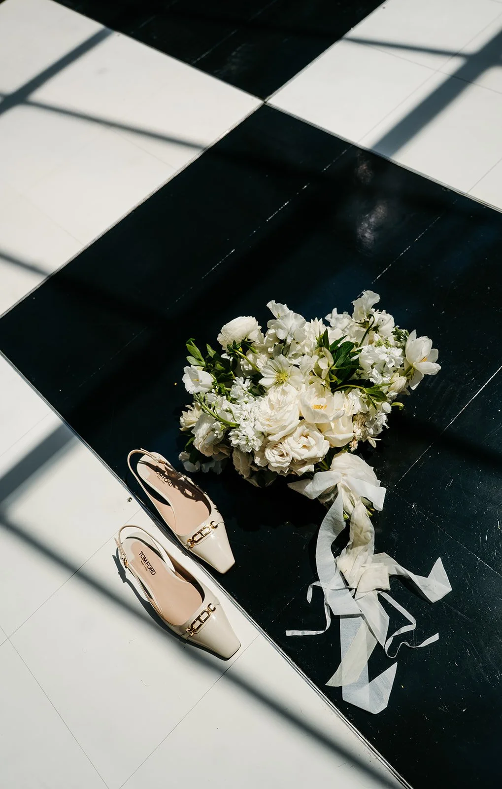 A bouquet of white flowers and a pair of cream-colored shoes on a black and white tiled floor.