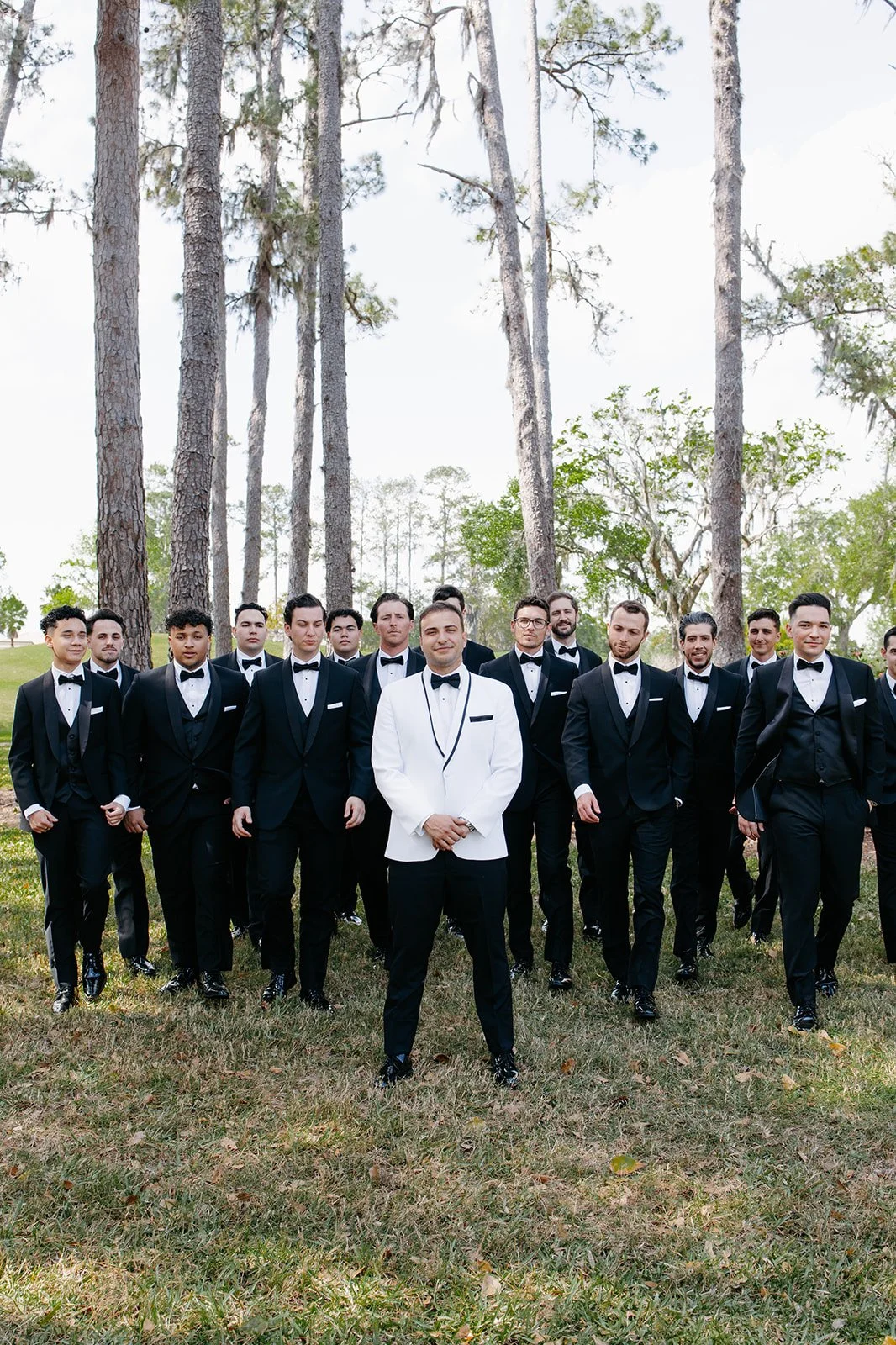 Group of men dressed in tuxedos outdoors, standing in front of tall trees during daylight.