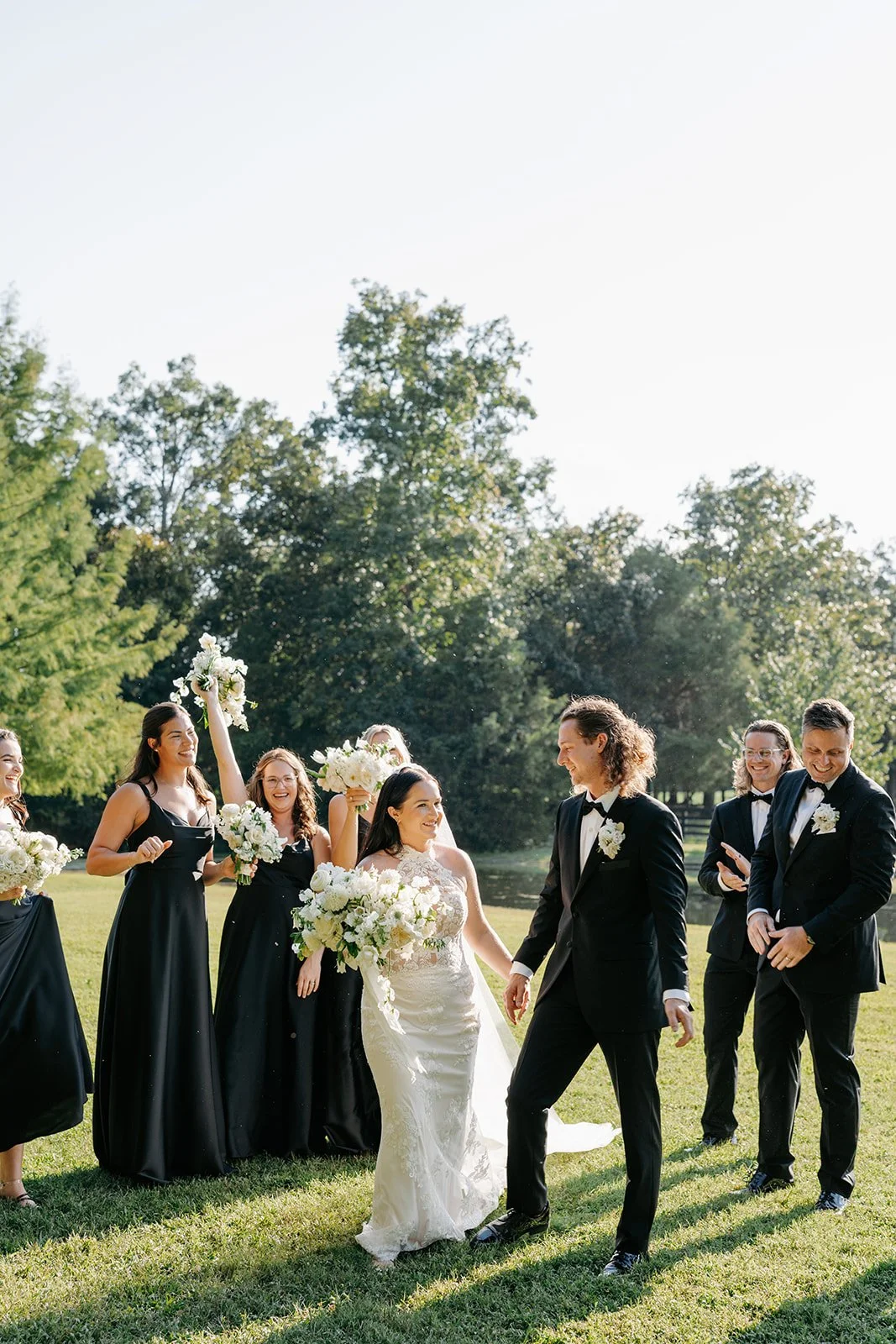 A bride and groom holding hands, walking outdoors on a wedding day, with bridesmaids and groomsmen celebrating around them in a lush green park.