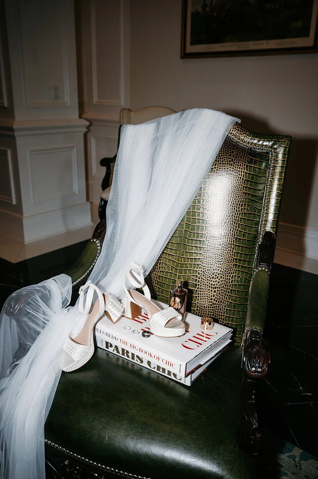 Green and gold crocodile-embossed armchair with items on the seat including white high heels, a white veil, a small perfume bottle, a gold ring, and two large books titled 'The Big Book of Chic Paris Chic' with a black and white cover.