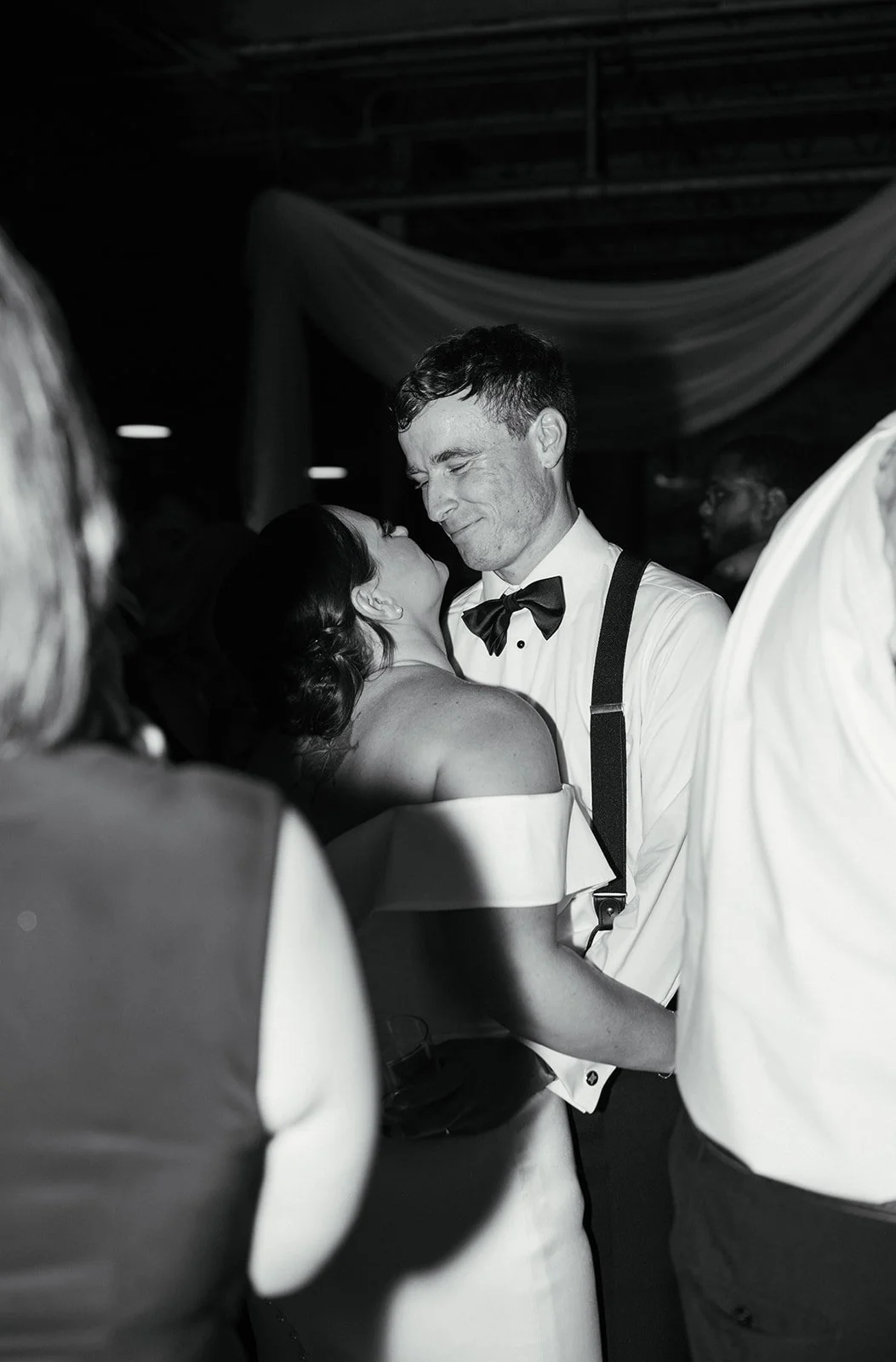 A black-and-white photo of a couple dancing closely at a wedding reception, surrounded by guests. The woman is wearing an off-shoulder dress, and the man is dressed in a suit with suspenders and a bow tie. They are gazing at each other lovingly.