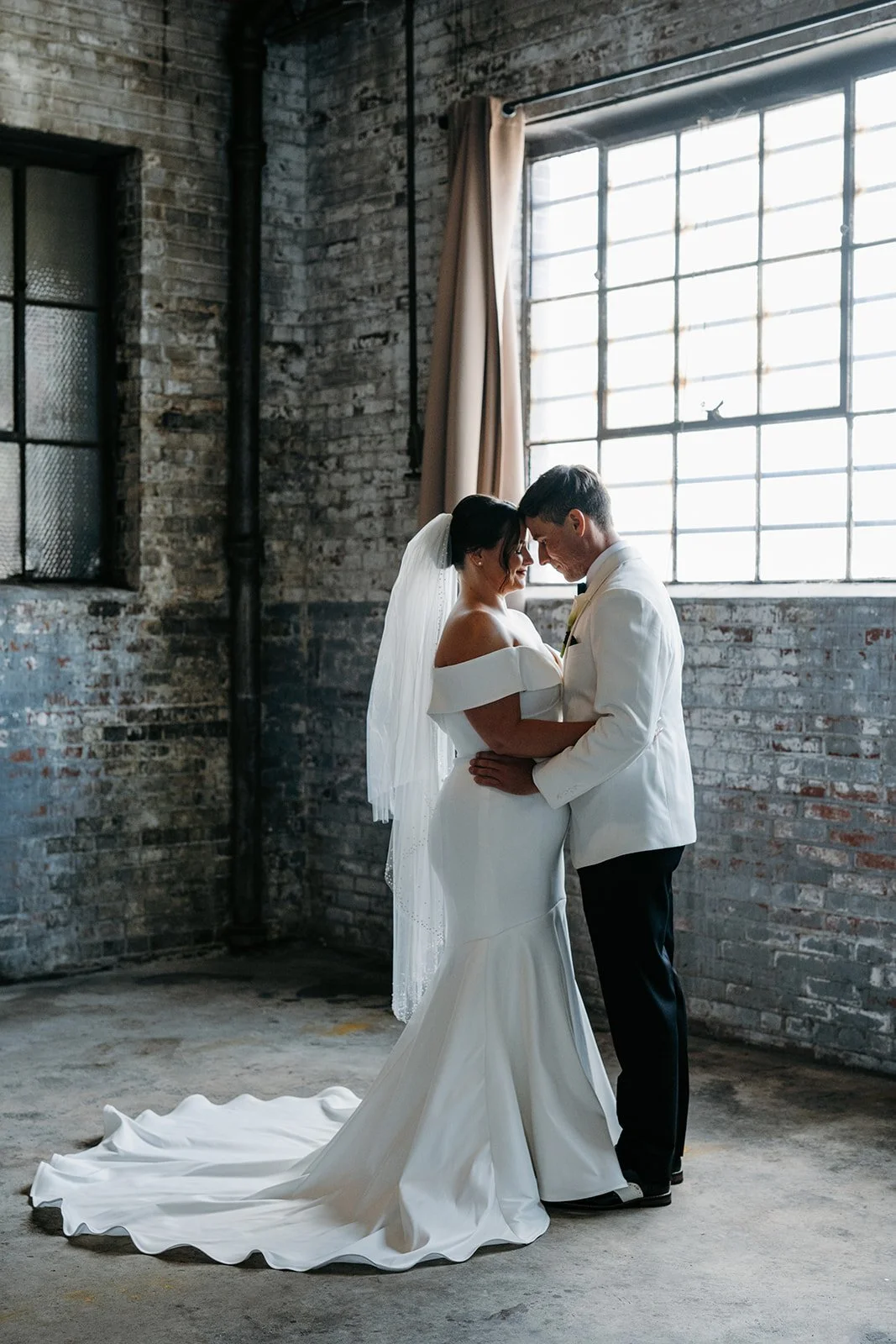 A bride and groom standing close together, touching foreheads, in an industrial-style room with brick walls and large windows.