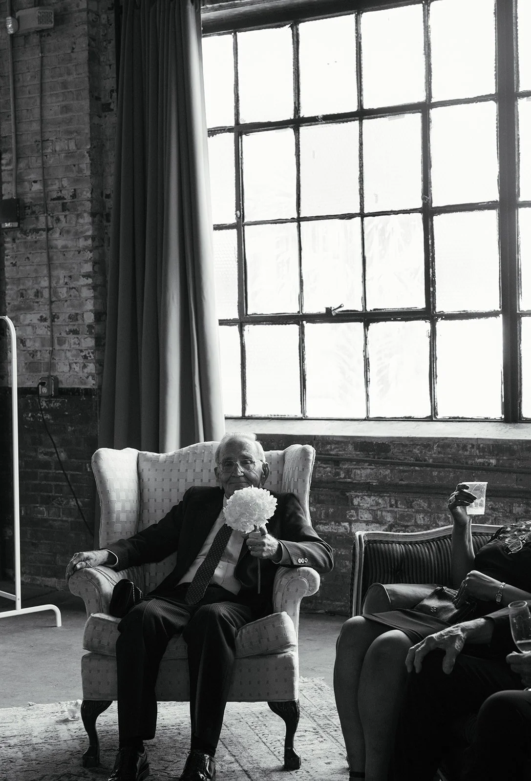 An elderly man in a tuxedo sitting in an armchair, holding a bouquet of flowers, in a room with large industrial-style window and brick walls.