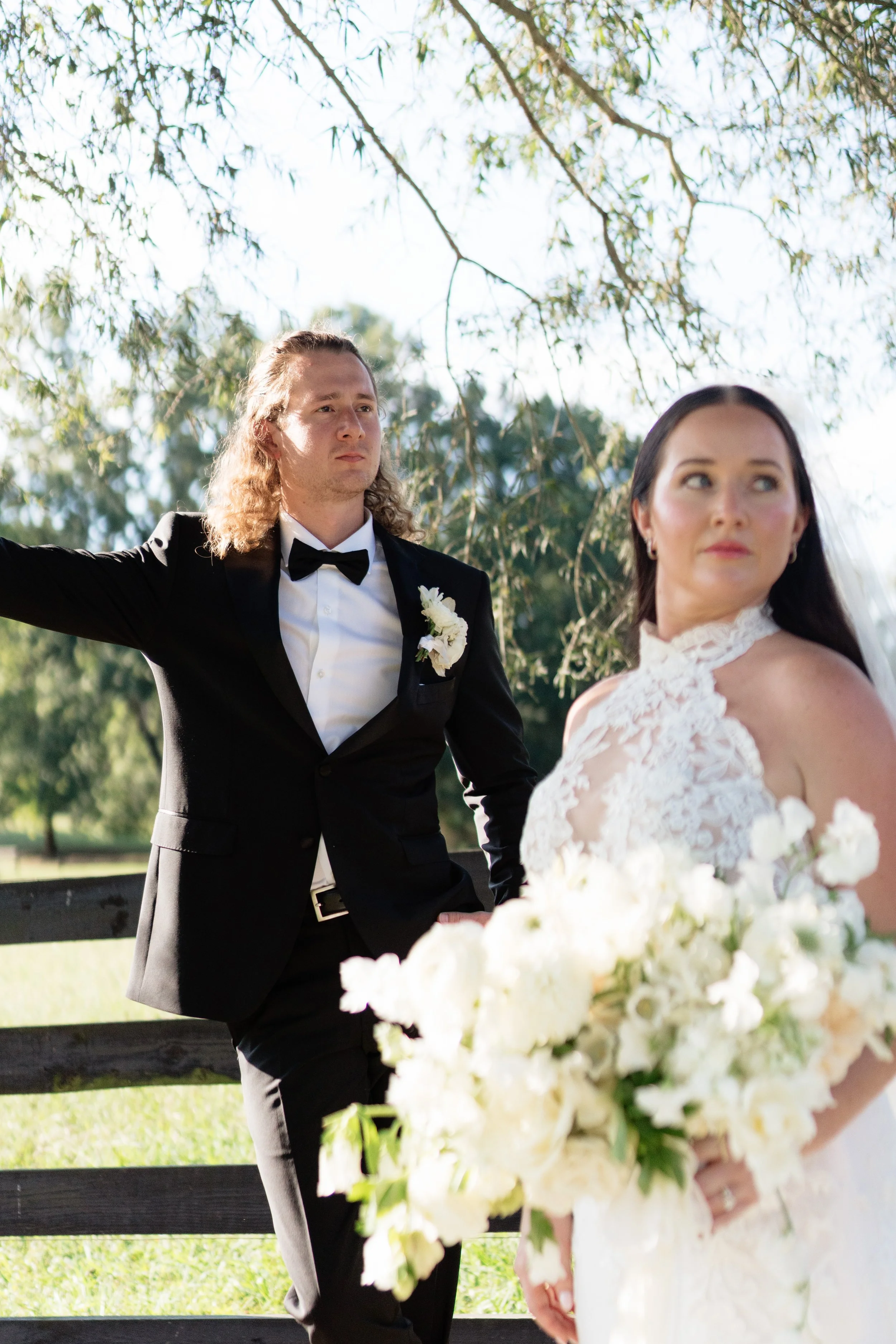 A bride holding a large bouquet of white flowers looking to the side and a groom in a black tuxedo with a white flower boutonniere standing outdoors under a tree, with a wooden fence and green grass in the background.