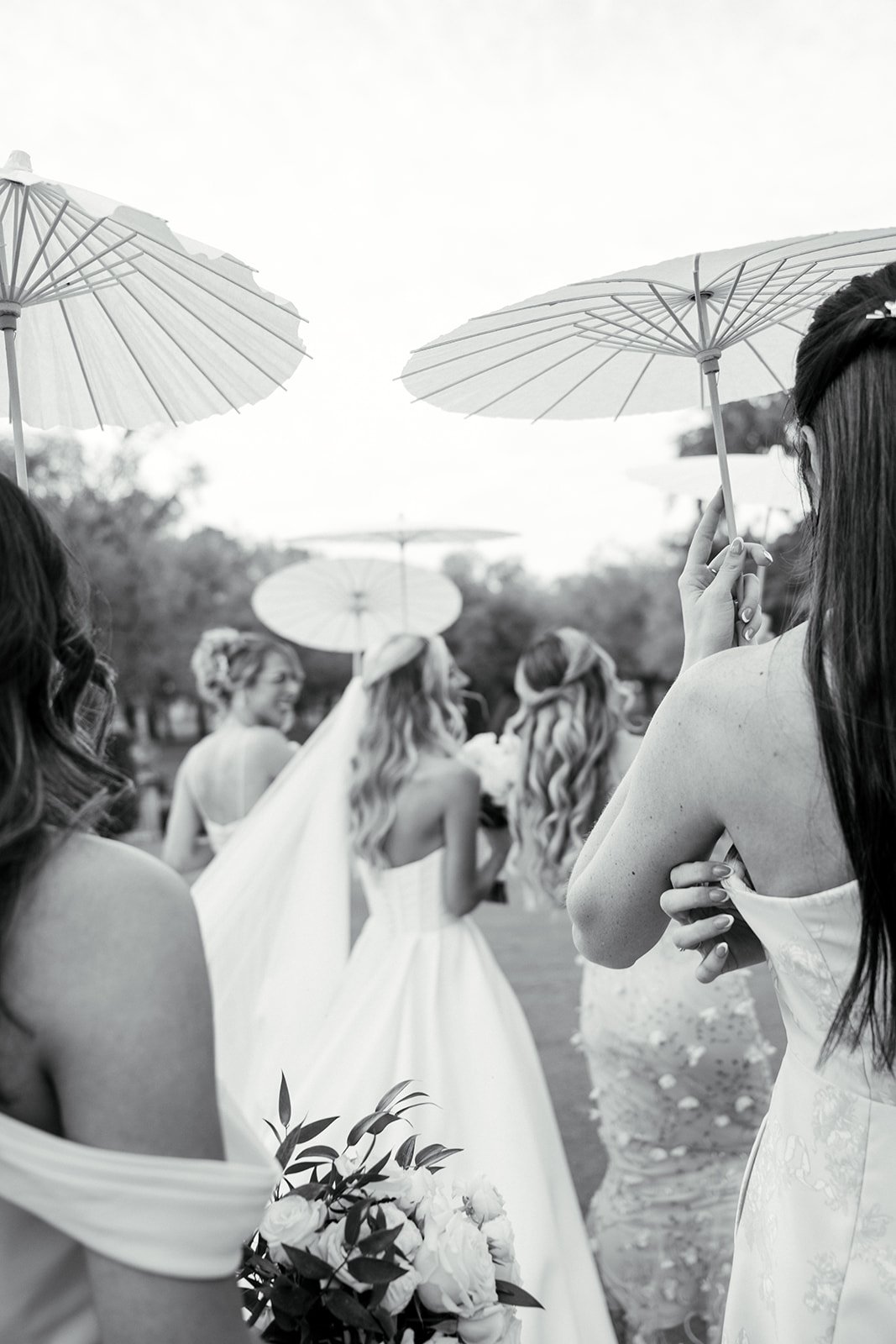 Group of women dressed in wedding attire, holding umbrellas, outdoors, black and white photo.
