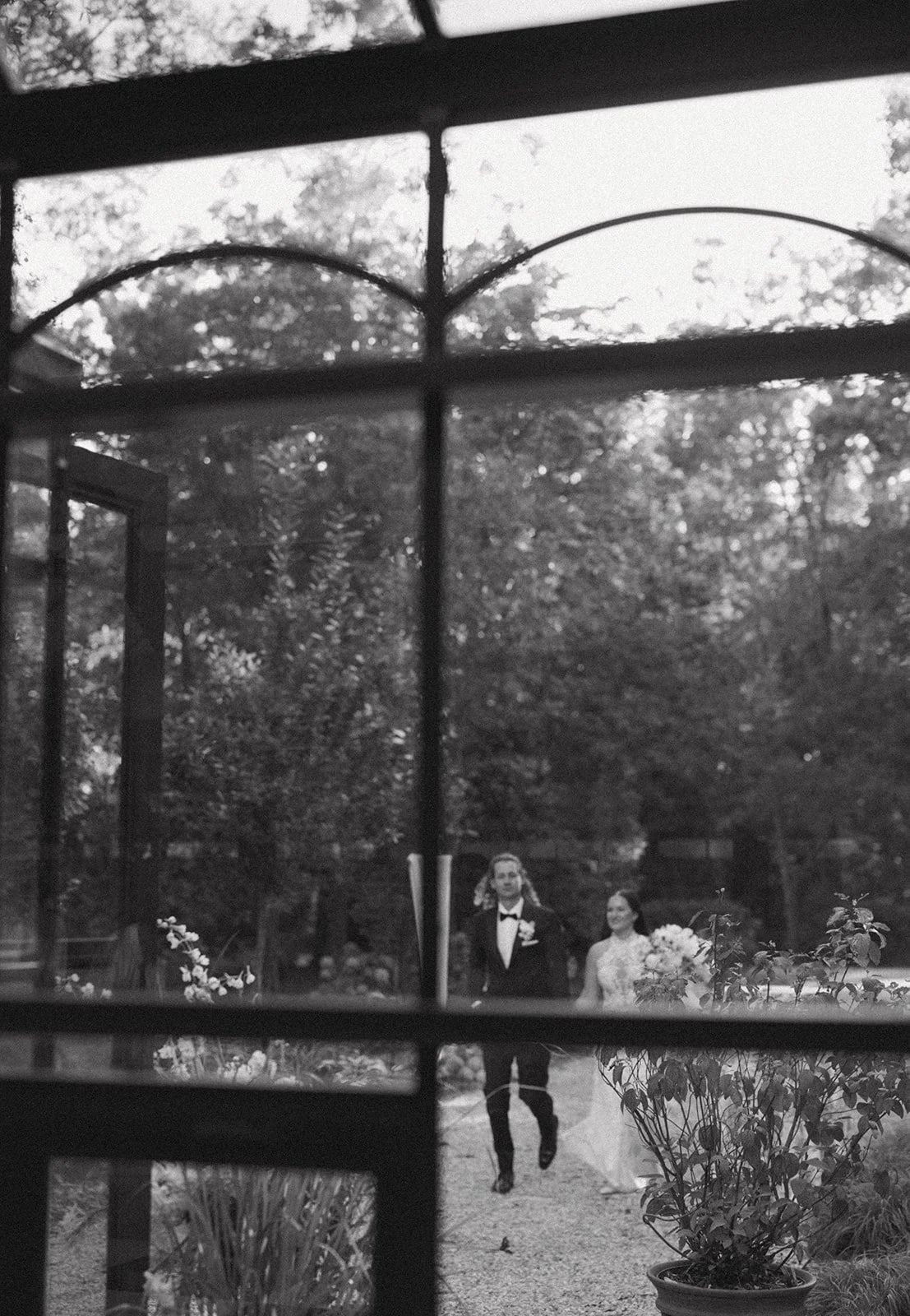 Black-and-white photo taken through a window with metal bars; a man and woman in formal attire walking outdoors in a garden with trees and plants.