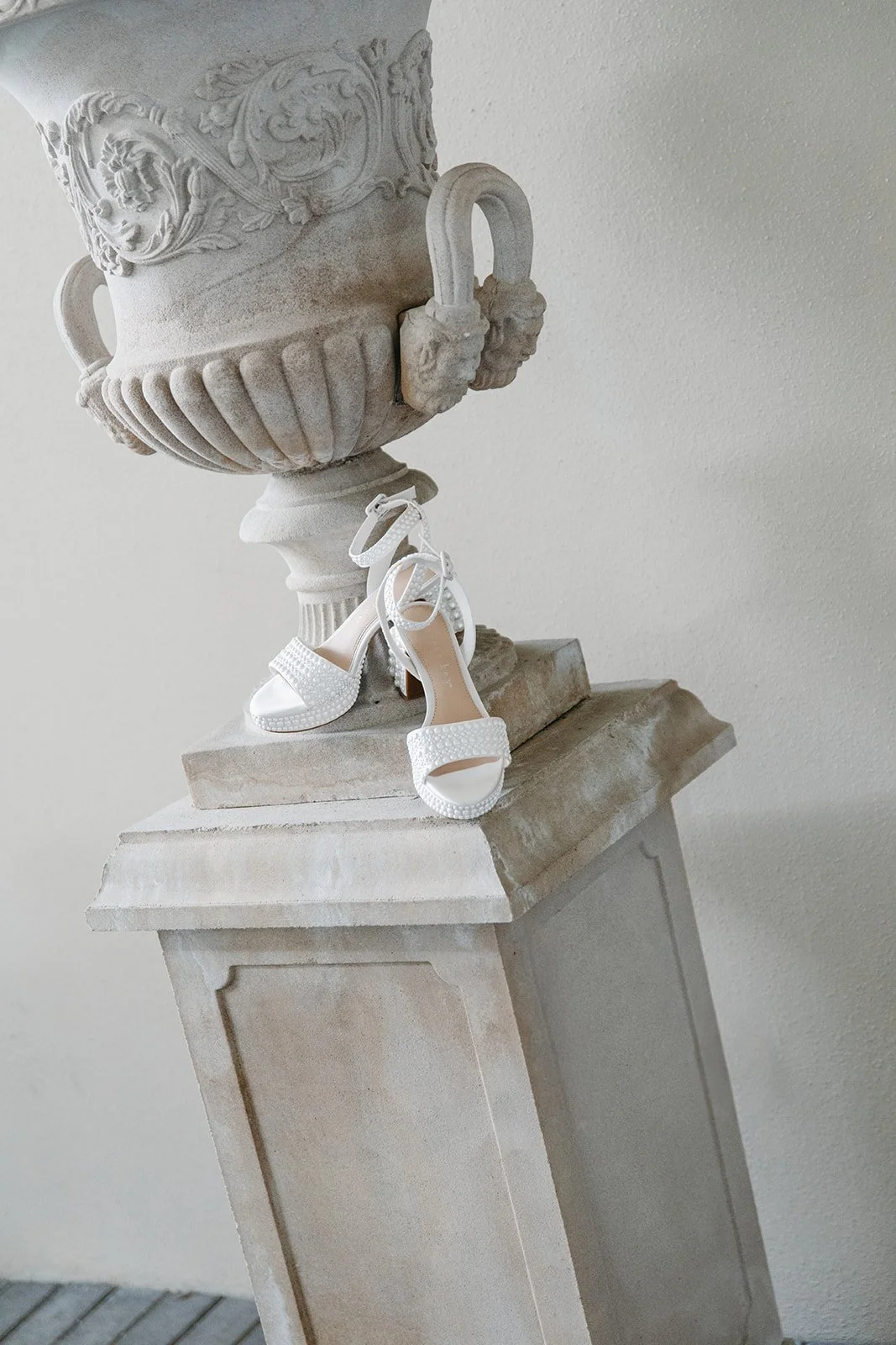White high-heeled sandals with ankle straps, displayed on a stone pedestal next to an ornate stone urn with lion head handles, outdoors against a plain wall.