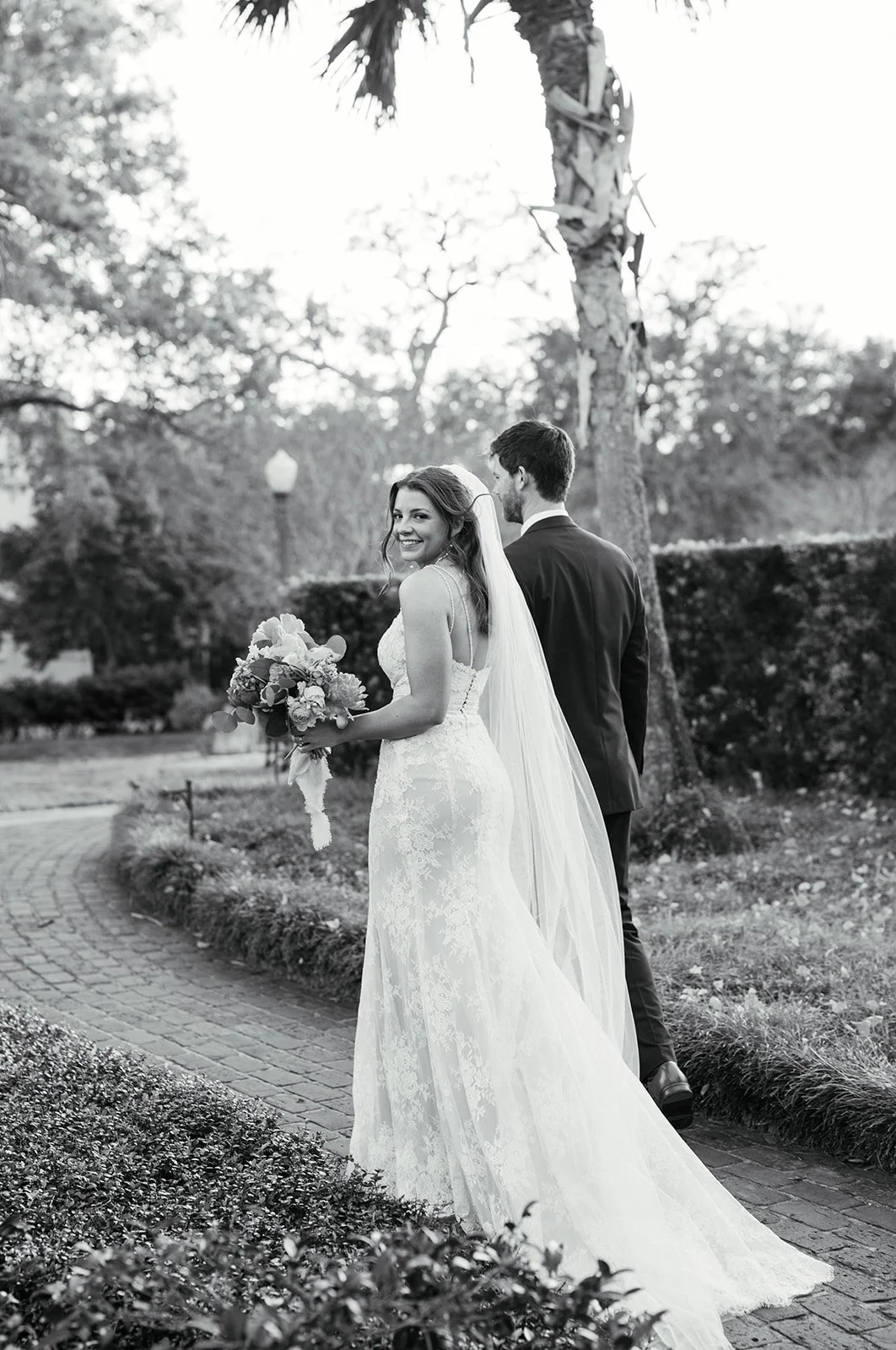 A bride in a lace wedding dress holding a bouquet of flowers, smiling at the camera, walking behind a groom in a suit, outdoors near trees and bushes in black and white.