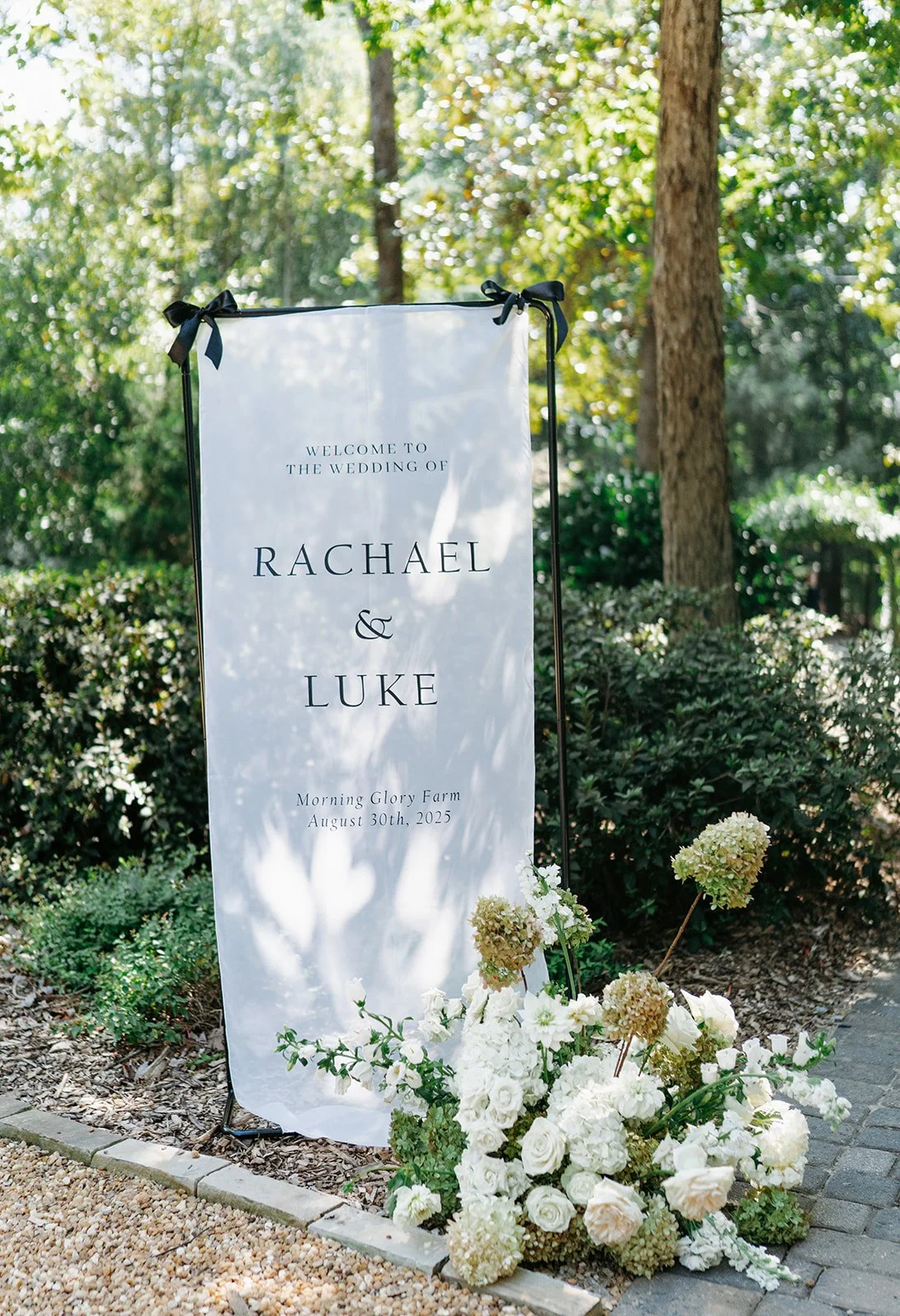 Outdoor wedding welcome sign for Rachael and Luke at Morning Glory Farm on August 30th, 2025, decorated with white flowers including roses and hydrangeas, with lush greenery and tall trees in the background.