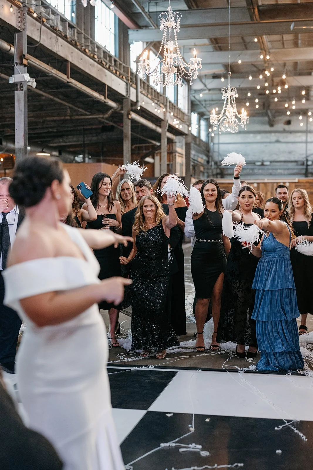 A bride in a white gown is giving a speech or toast at her wedding reception, with guests in formal dresses and suits watching her, some holding feathered noise makers in a decorated industrial-style venue with chandeliers.