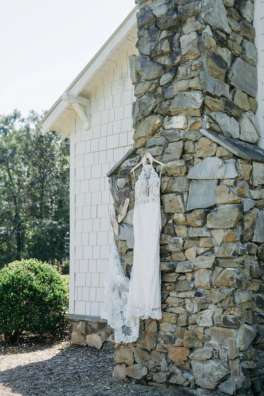 A white lace wedding dress hanging on a wooden hanger, next to beige high-heeled shoes, hanging on a stone chimney outside of a house with white siding.