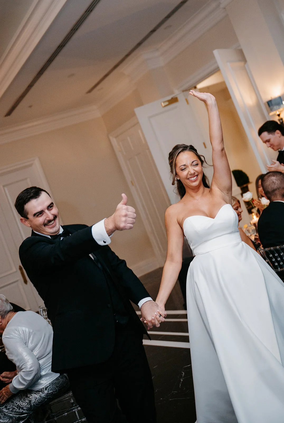 A woman and a man in wedding attire holding hands and celebrating at a wedding reception, with others in the background.