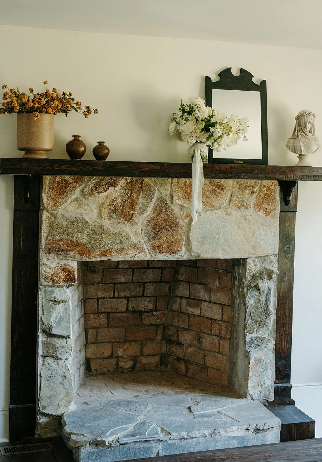 A rustic fireplace with a stone and brick interior, topped with a dark wood mantel. Decor includes a large beige vase with yellow flowers, two small brown vases, a black mirror, a white bust sculpture, and a floral arrangement with white flowers and 