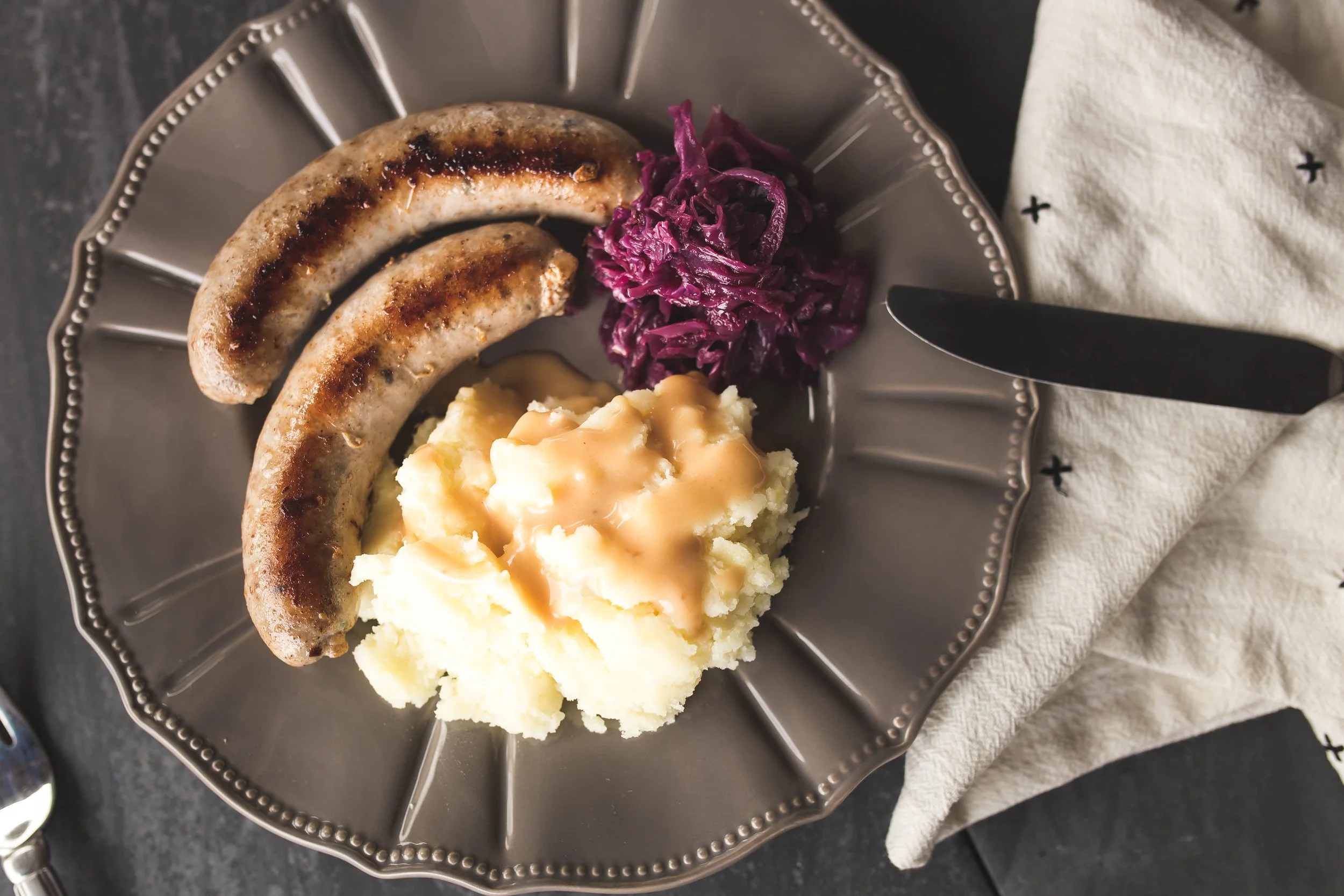 Two grilled sausages on a plate with cooked cabbage and mushed potatoes with gravy.
