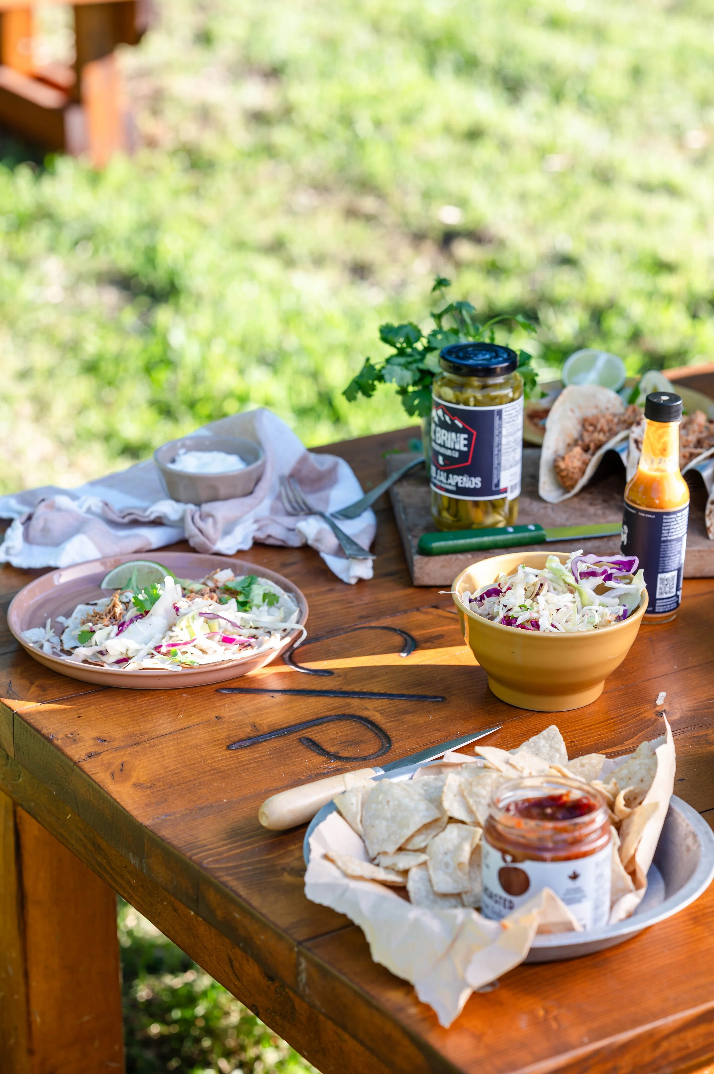 a DYI taco bar set up on a picnic table, includes hot sauce, chips and salsa, and coleslaw