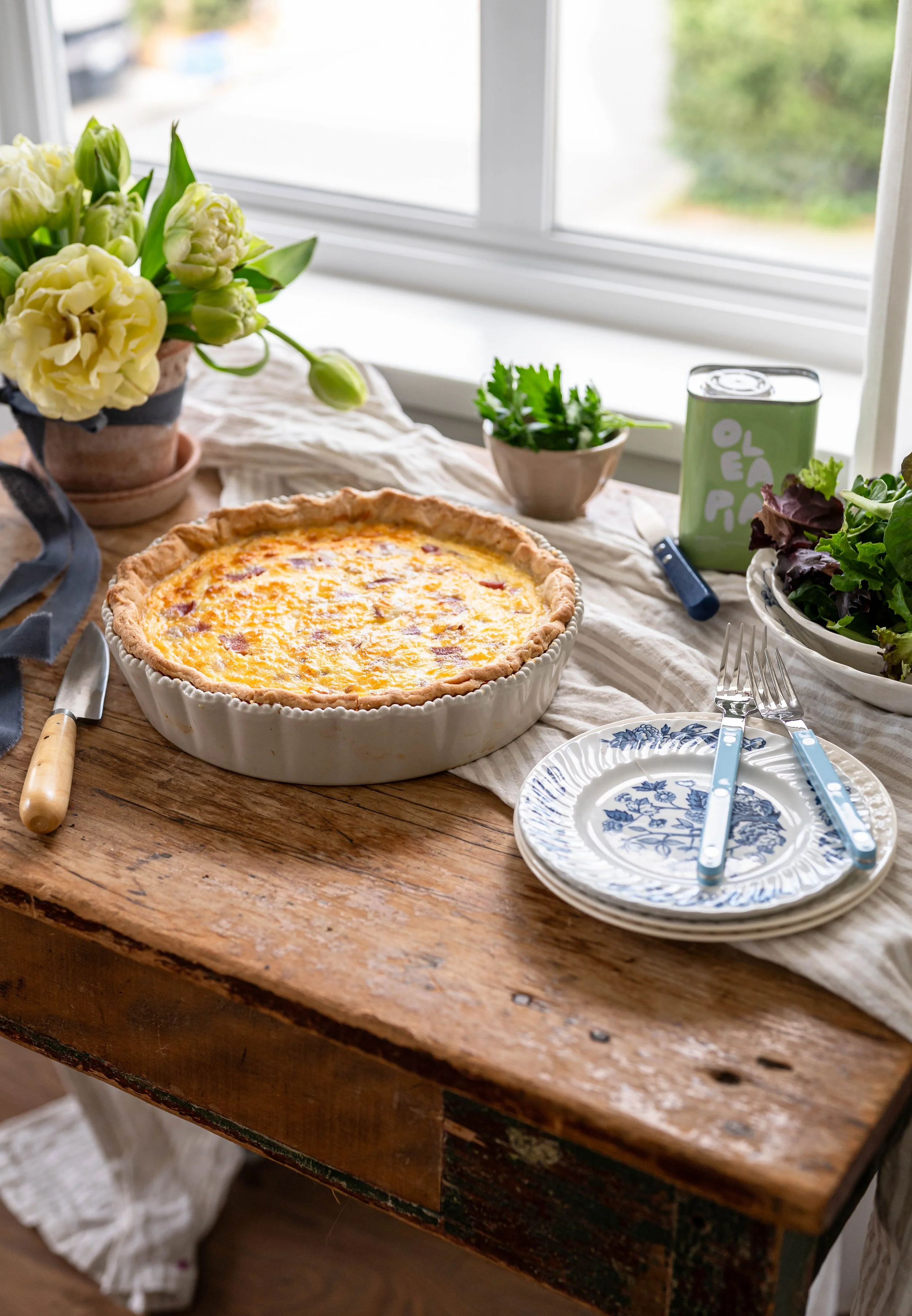A rustic wood table with a classic quiche, a stack of blue plates and tulips