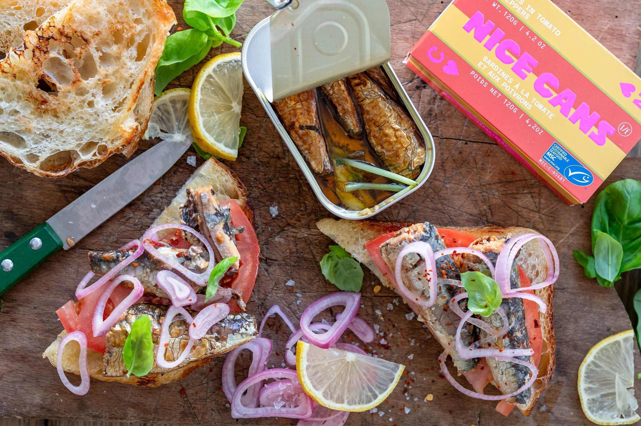 A cutting board with a brightly coloured package that says Nice Cans and a can of sardines opened with sardine toast