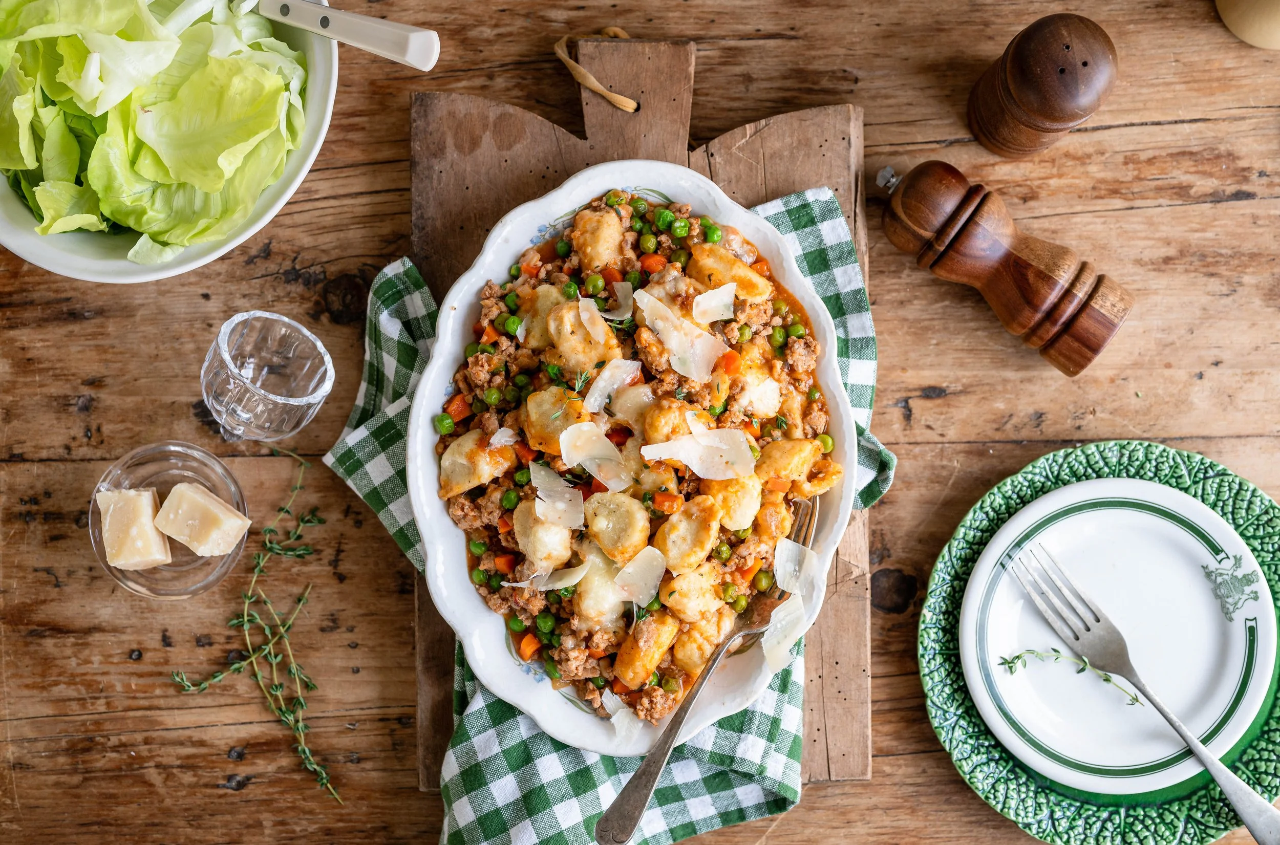 A white plate of ground beef, gnocchi, and vegetables covered in grated parmesan. It's sitting on a wooden table with a bowl of fresh salad