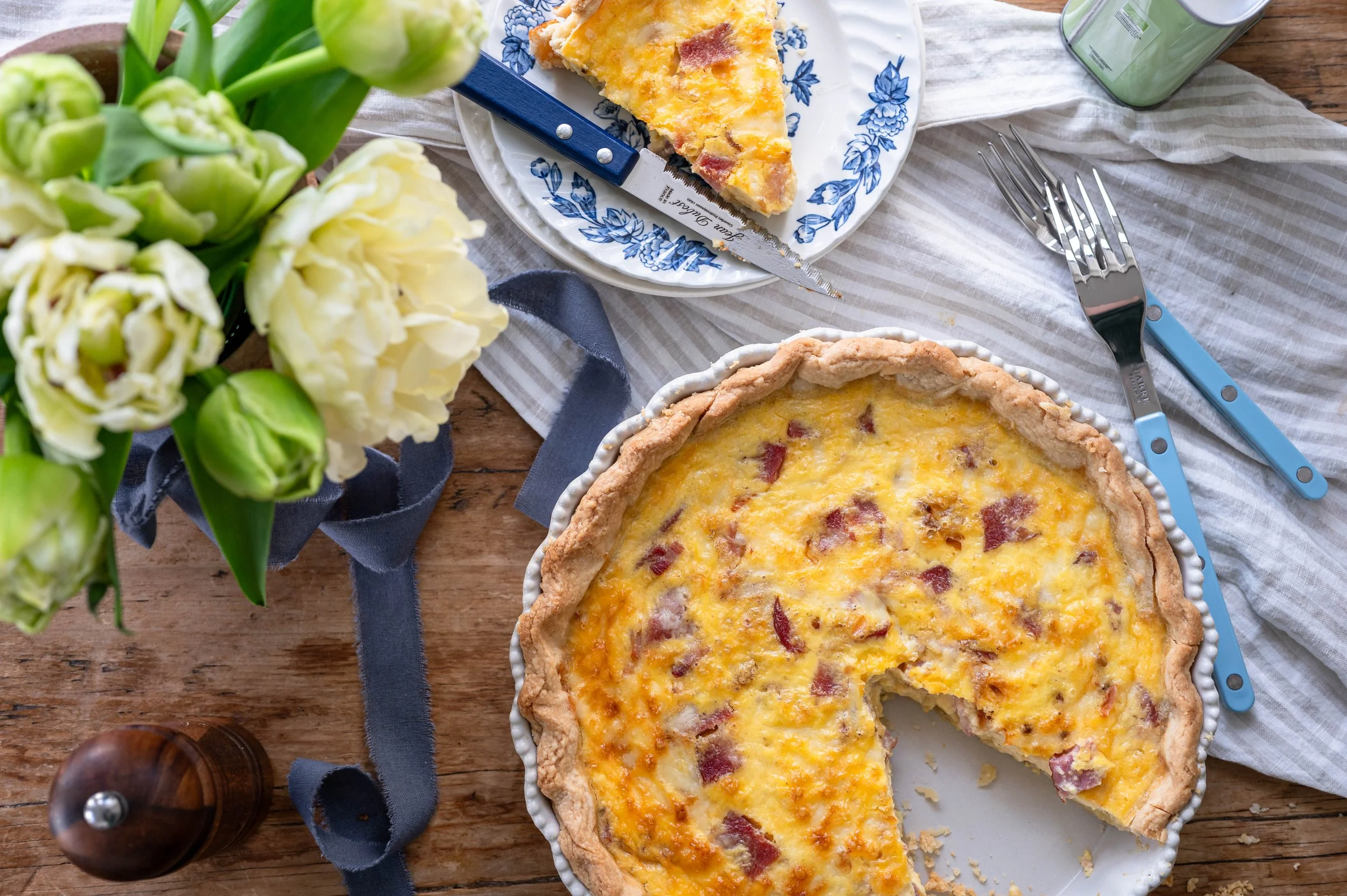 An egg and ham quiche with a slice removed on a table with tulips and a stripped table cloth