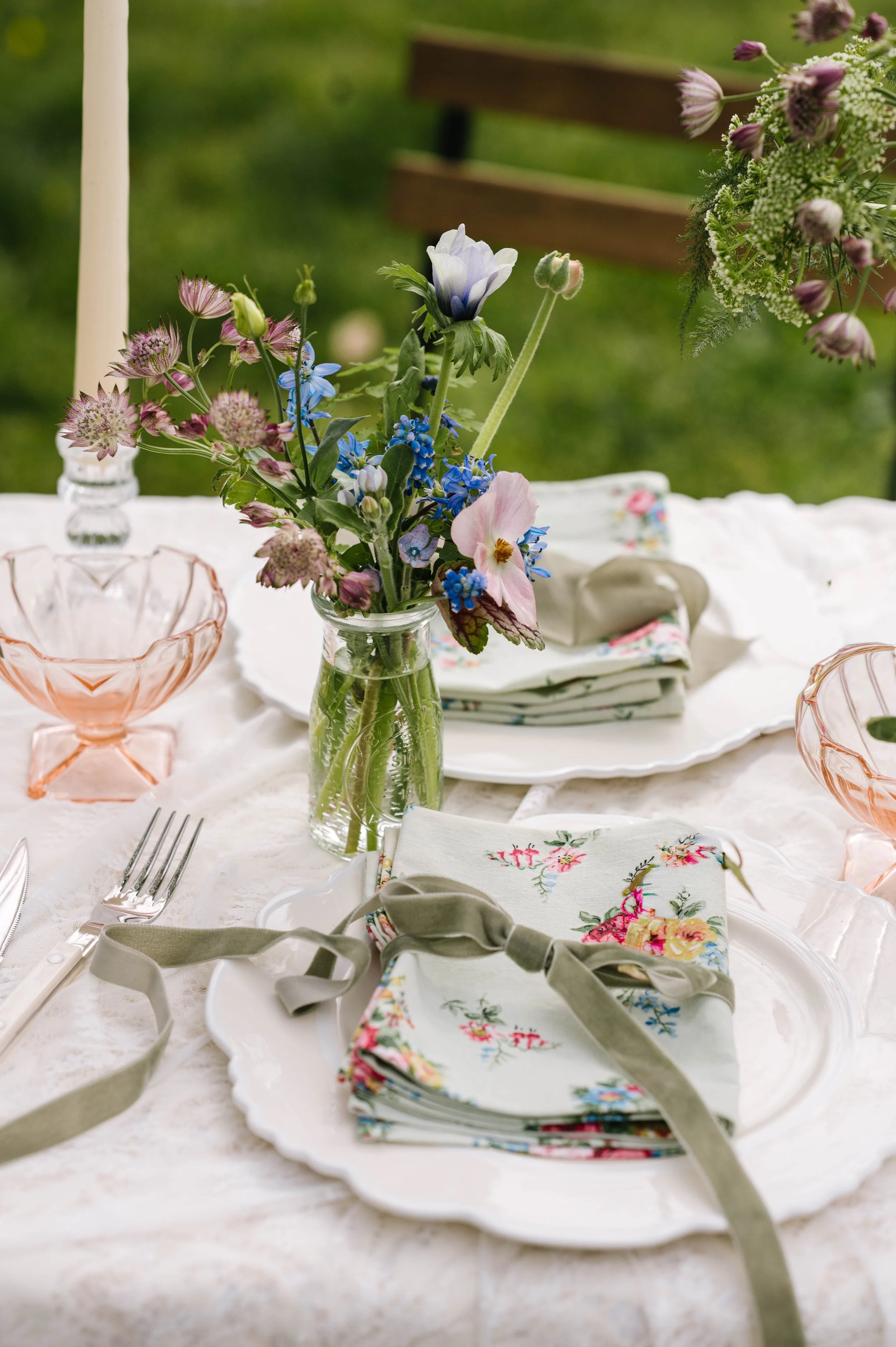 A table set for Mothers Day with a lace table cloth, bud vases with pink and blue flowers and floral napkins and taper candles
