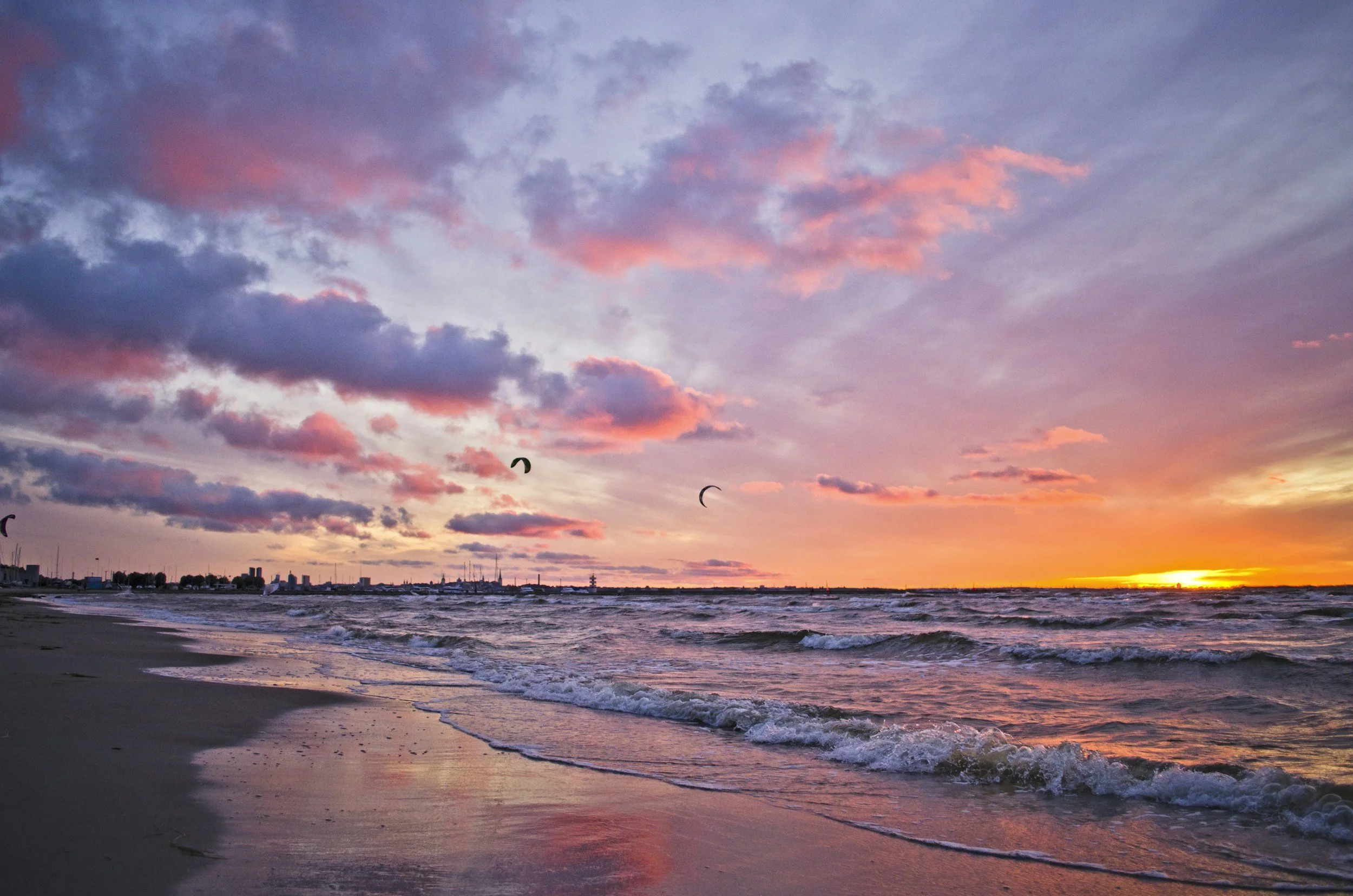 A sunset over the ocean with pink and purple clouds and a few kite surfers in the distance.