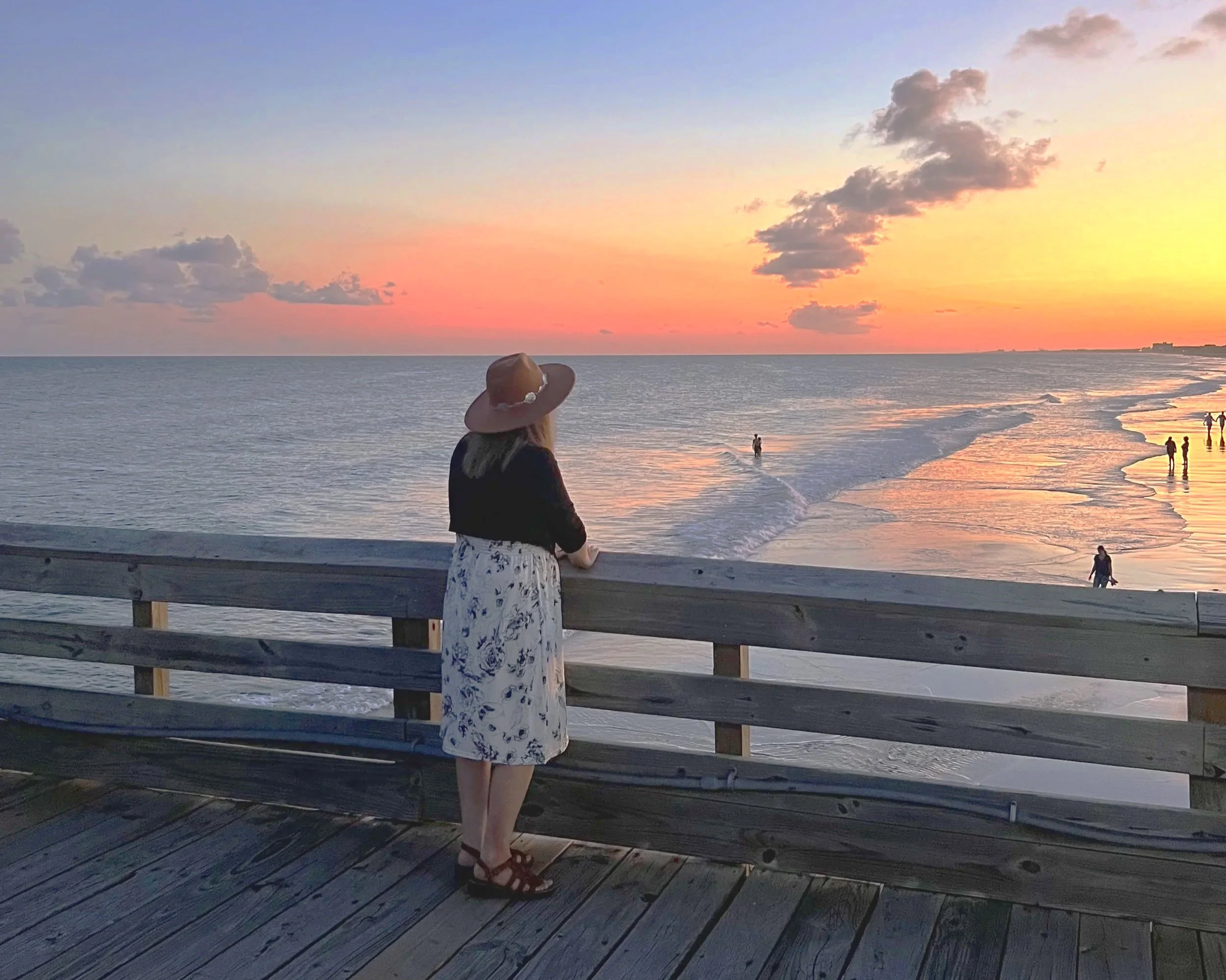 Me with blonde hair wearing a wide-brimmed hat and a black and white floral dress stands on a wooden pier, gazing at the sunset at the beach.